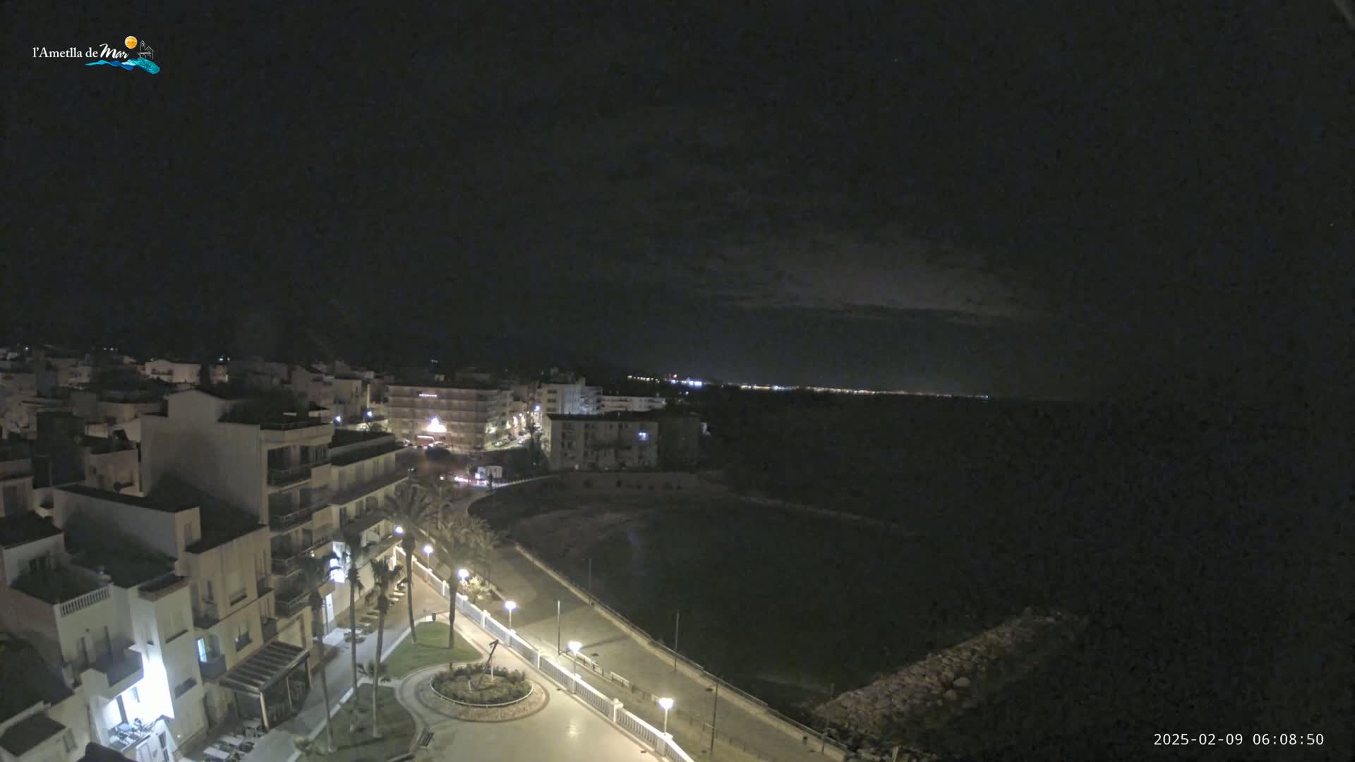 A nighttime, high-angle view shows a coastal town with buildings illuminated, a dark body of water, and a paved walkway lined with palm trees under a mostly clear sky.