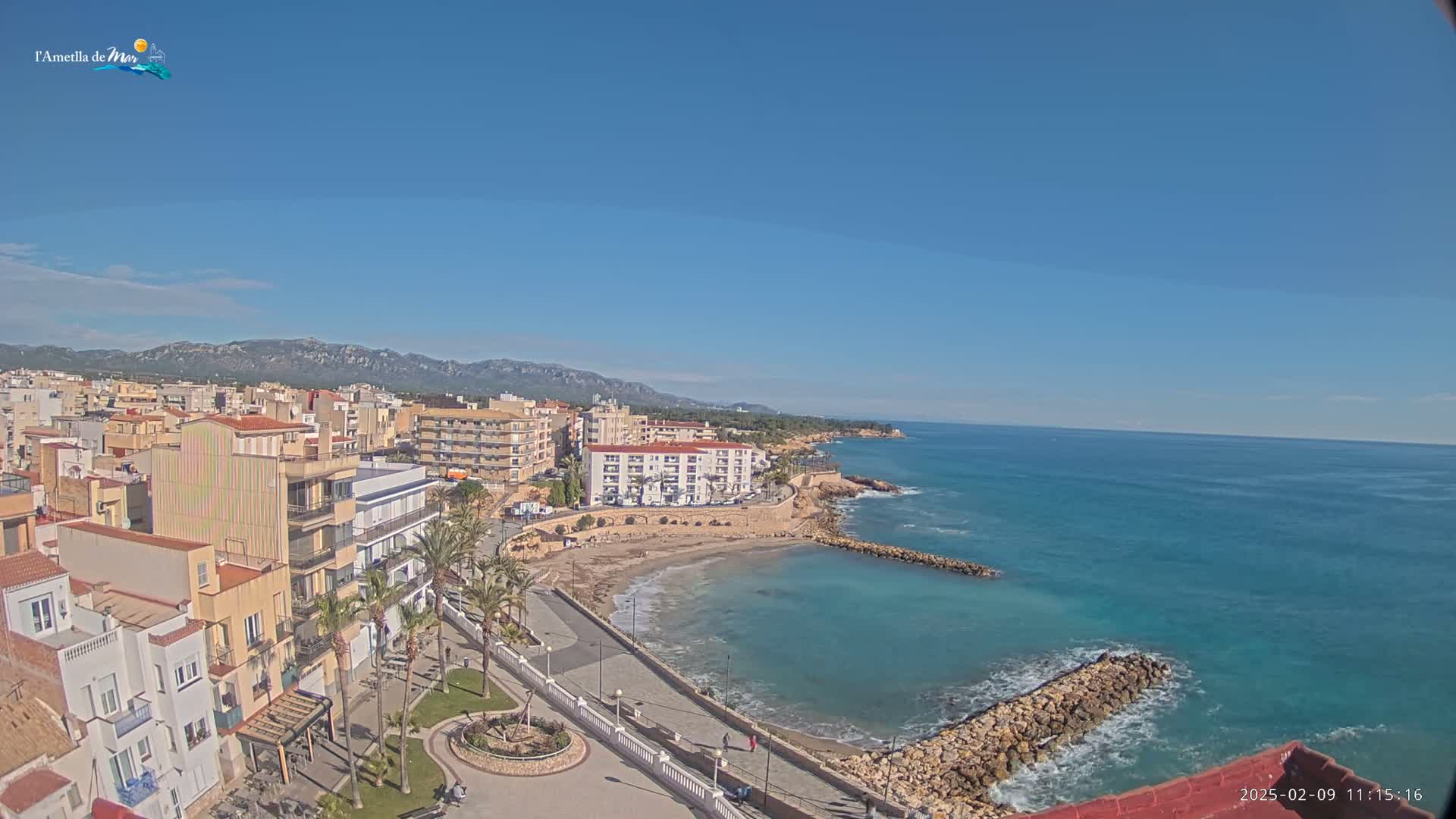 A coastal town with buildings, palm trees, and a sandy beach is visible on a clear, sunny day, with calm, turquoise water extending to a mountain range in the distance.