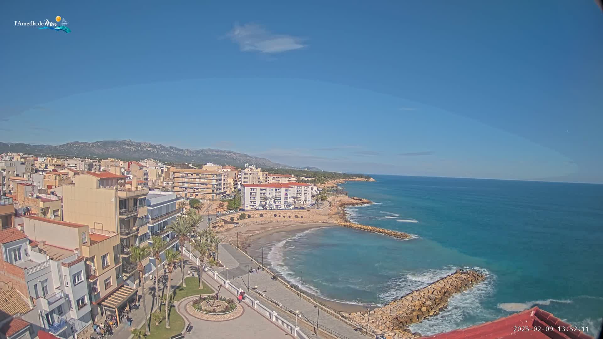 A coastal town with buildings, palm trees, and a sandy beach is visible on a clear, sunny day, next to a calm, blue ocean with small waves.