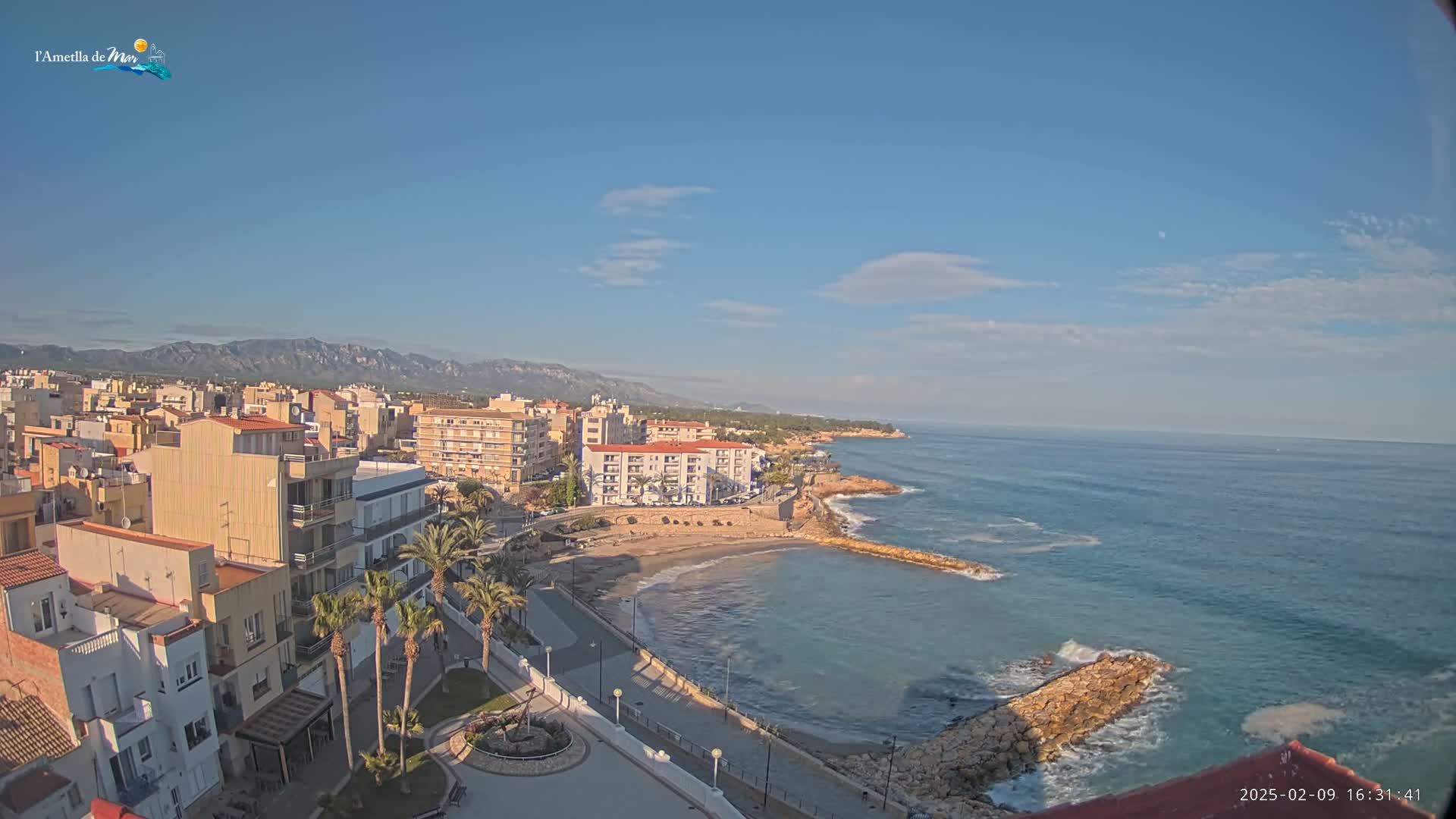 A coastal town with buildings and palm trees is situated next to a calm ocean under a mostly clear, sunny sky with a mountain range visible in the distance.