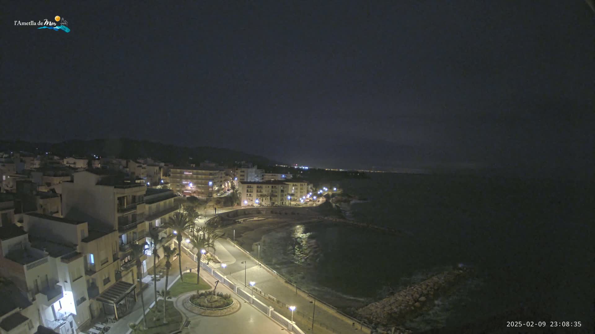A nighttime coastal town with a paved waterfront path and buildings illuminated by streetlights, under a clear dark sky.