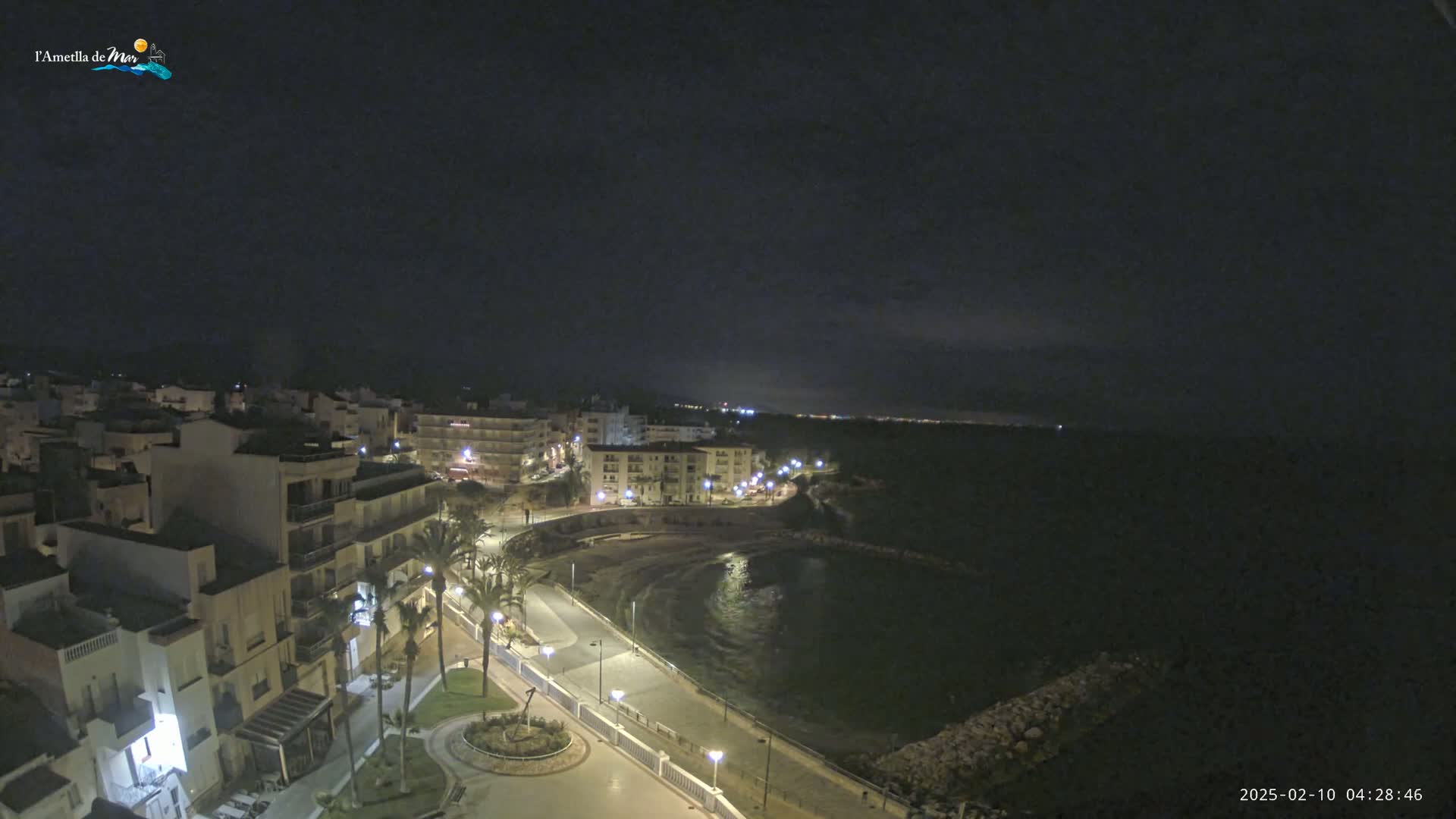 A nighttime, high-angle view shows a coastal town with its lights on, a calm bay, and a paved walkway along the shoreline.