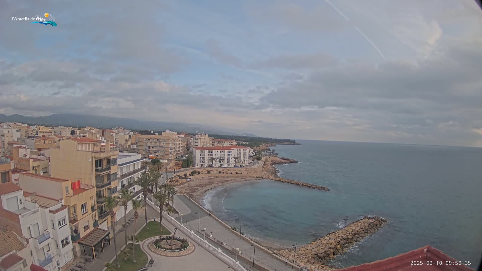 An aerial view shows a coastal town with buildings along a calm, blue sea under a mostly cloudy sky.