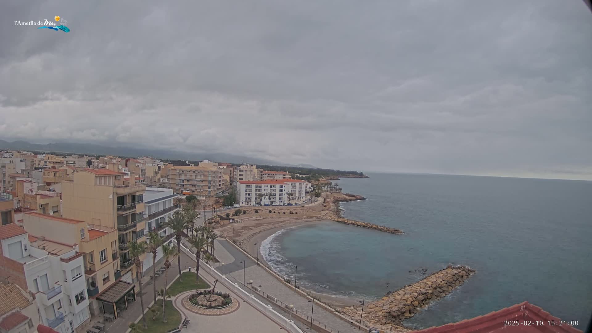 An overcast day reveals a coastal town with buildings, palm trees, a small beach, and a calm sea.