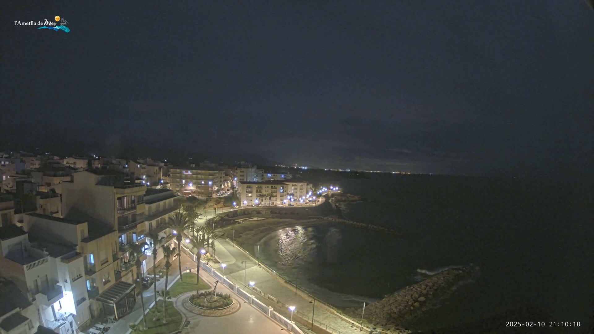 A nighttime, high-angle view shows a coastal town with buildings lit up along a paved waterfront path next to a calm, dark-colored body of water.