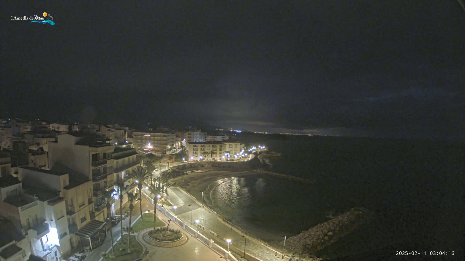 A nighttime coastal town scene shows a crescent-shaped bay with a paved walkway, buildings, and palm trees under a dark, cloudy sky.