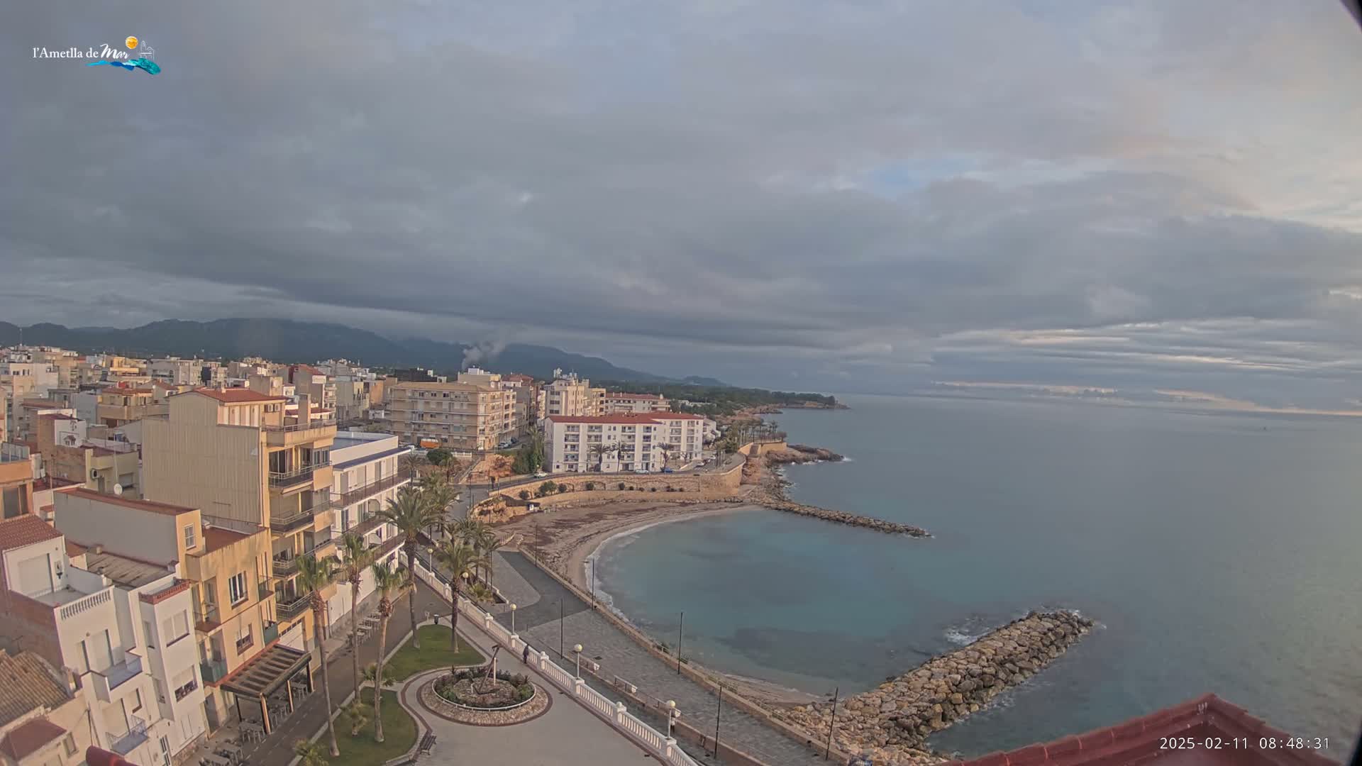 A coastal town with buildings and a small beach is seen under an overcast sky, with calm water extending to the horizon.