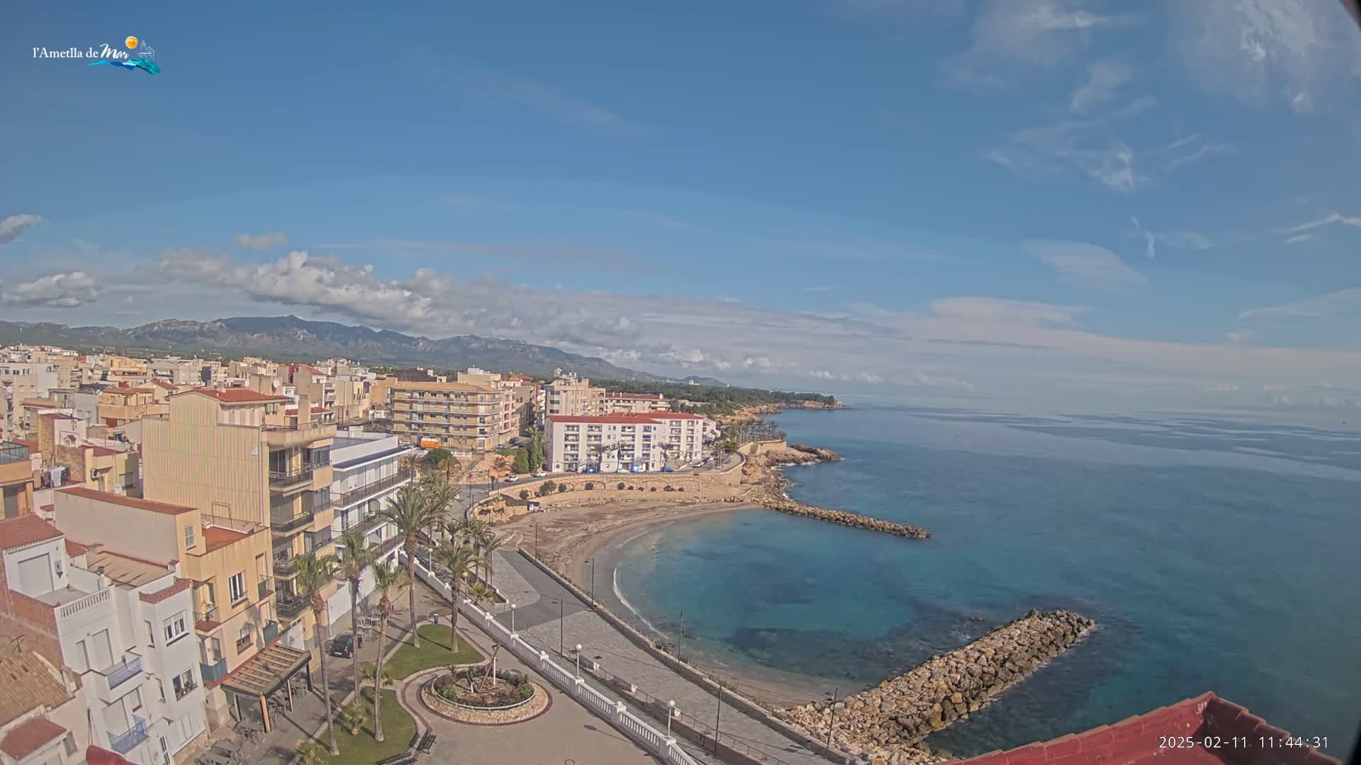 A coastal town with multi-story buildings, a small sandy cove, and a calm blue sea under a mostly sunny sky with some scattered clouds.