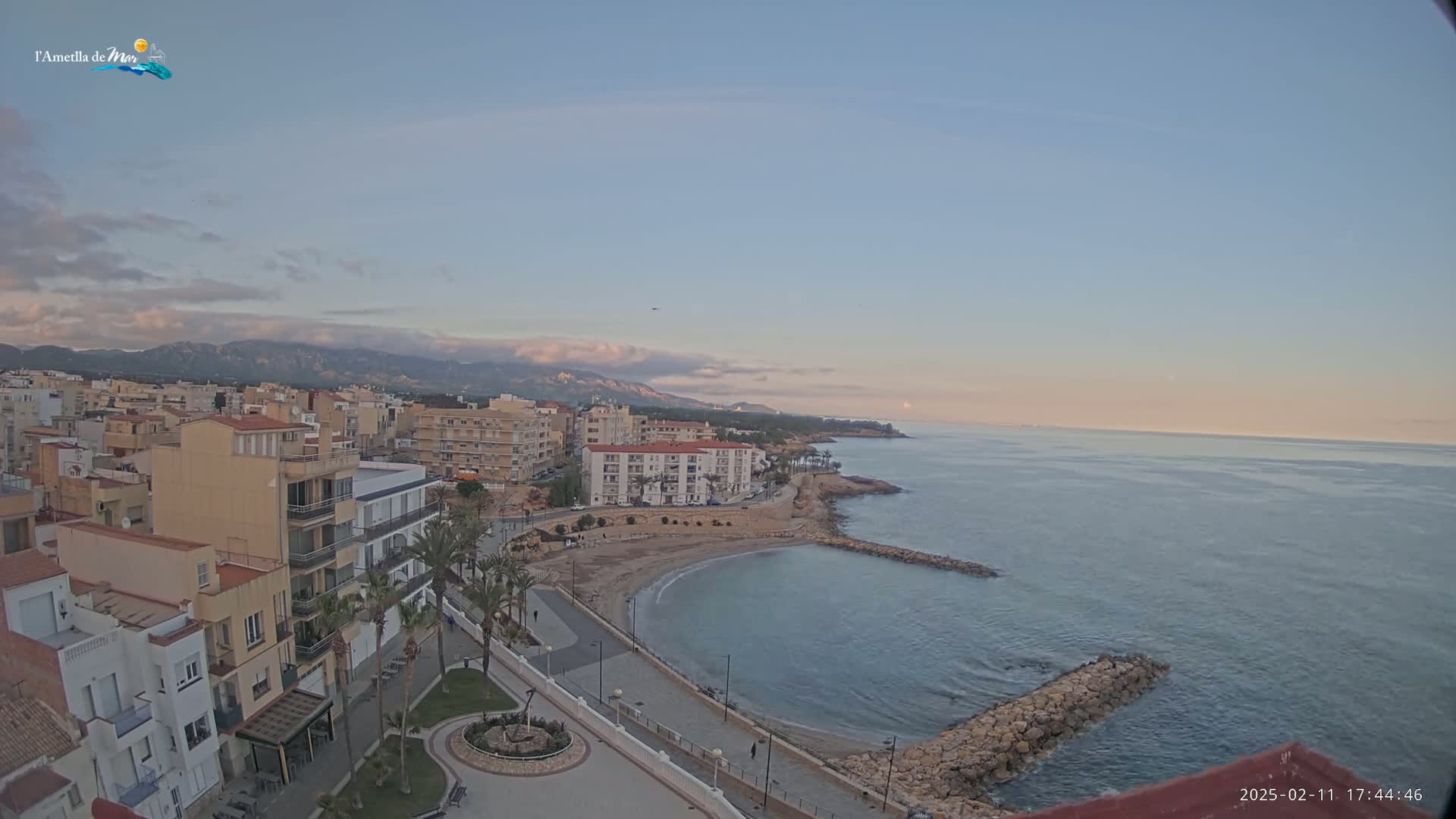 A coastal town with buildings and palm trees is situated next to a calm sea under a partly cloudy sunset sky.