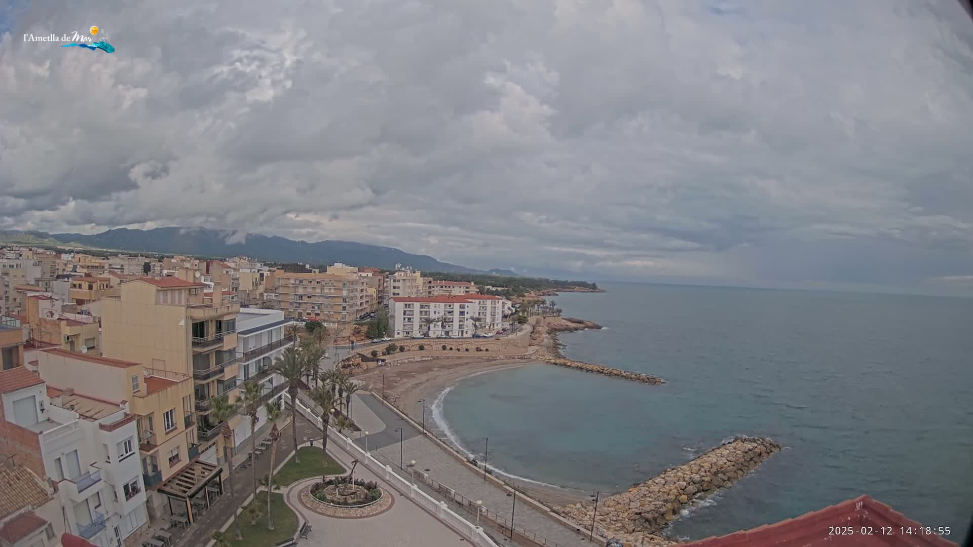 An overcast sky hangs over a coastal town with buildings lining a curved bay, where a small beach and a rocky breakwater are visible.