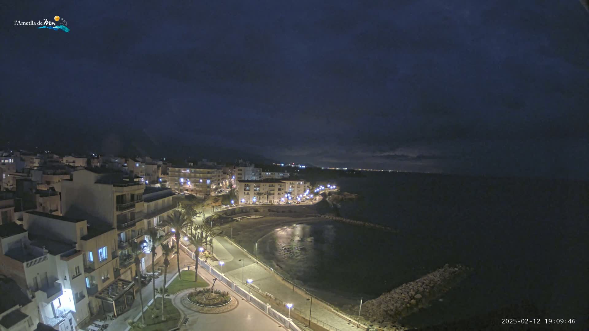 A nighttime coastal town is illuminated, with dark clouds overhead and a calm, dark sea stretching to the horizon.