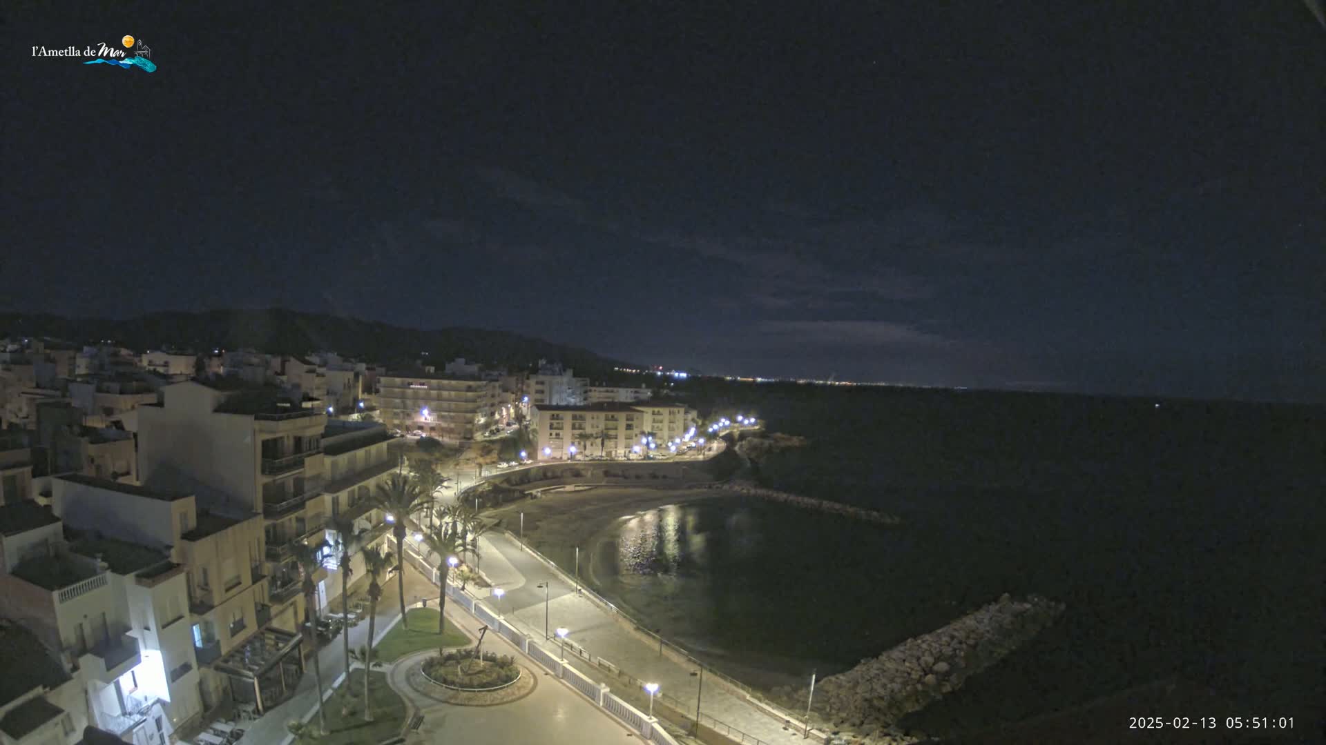 A nighttime aerial view shows a coastal town with buildings lit up along a crescent-shaped bay under a dark, mostly clear sky.