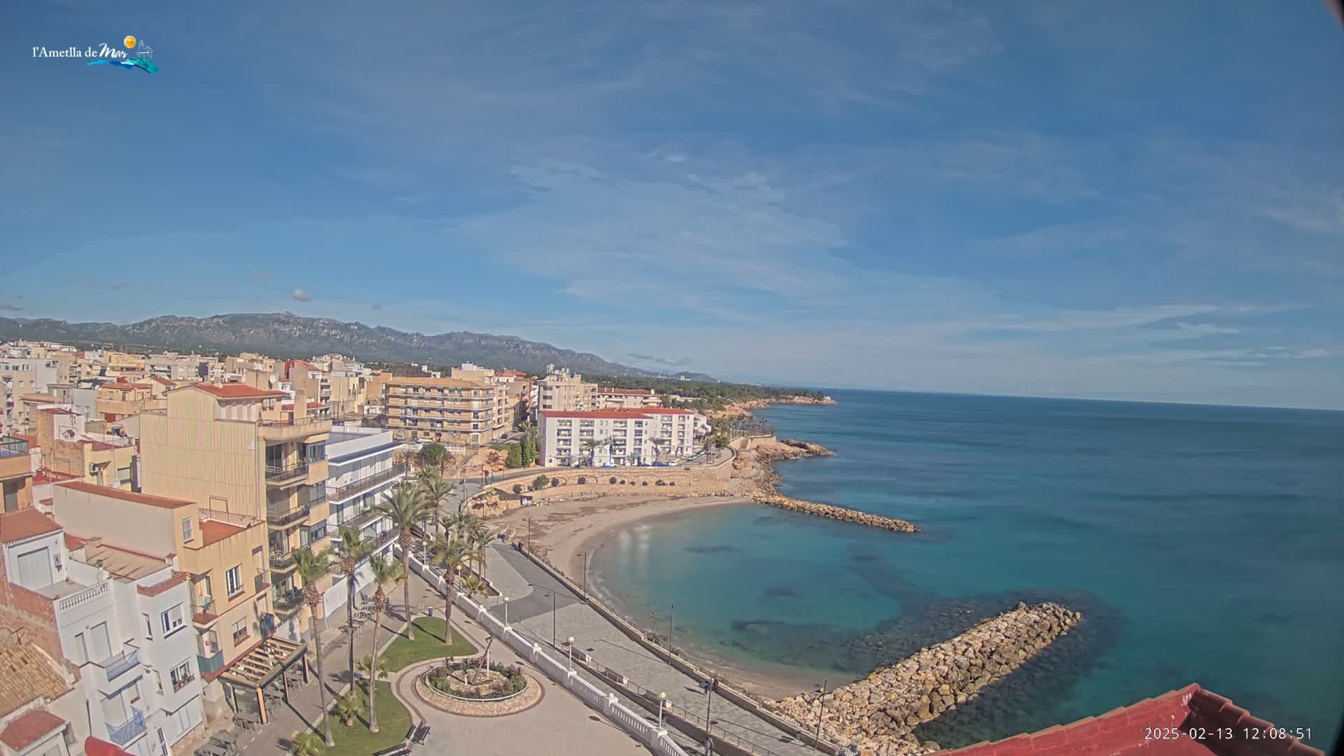 A coastal town with buildings, palm trees, a small sandy beach, and a calm blue sea under a mostly clear sky is shown from an elevated viewpoint.