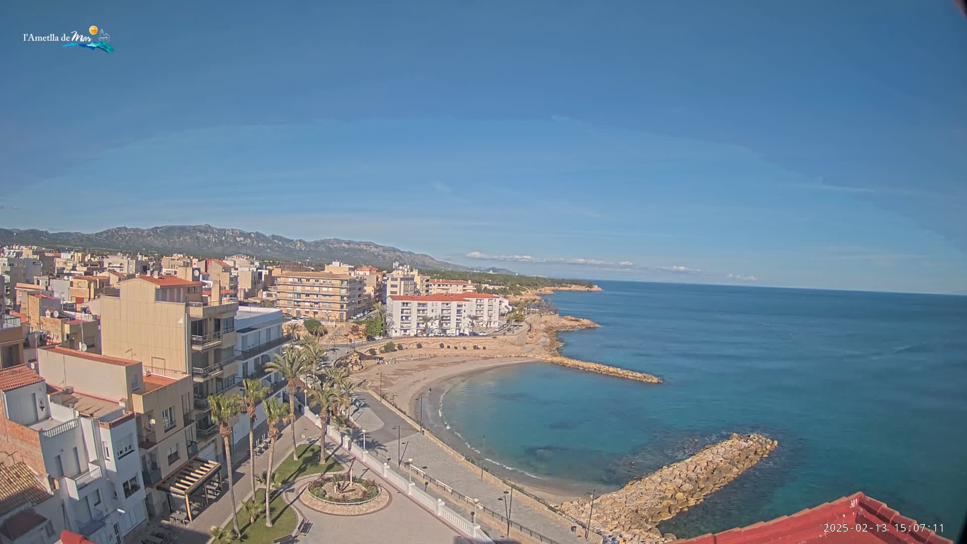 A coastal town with buildings and palm trees sits on a small, sandy beach, under a clear blue sky, next to a calm, teal ocean with a rocky shoreline.