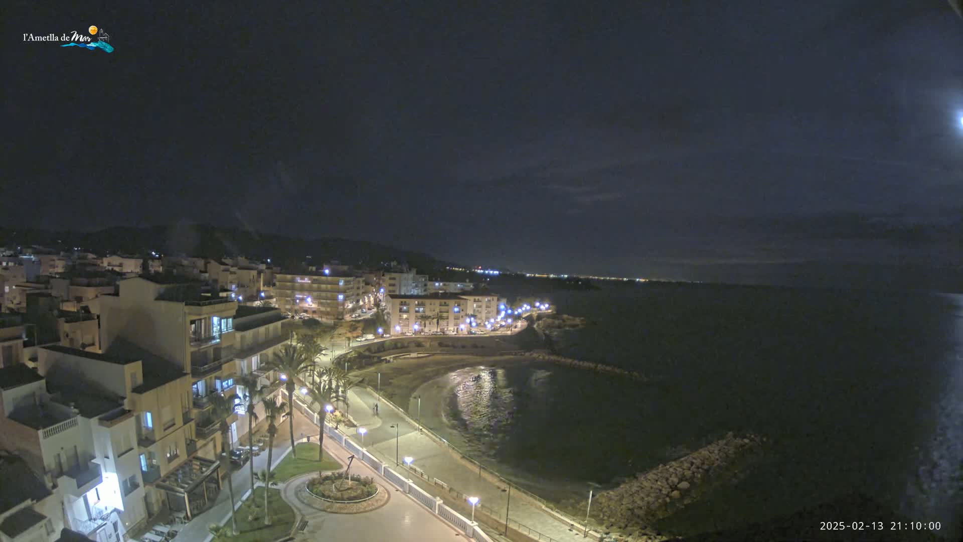 A nighttime coastal town scene shows a crescent-shaped bay with a paved walkway lined with lights, buildings, and palm trees, under a mostly clear, dark sky.