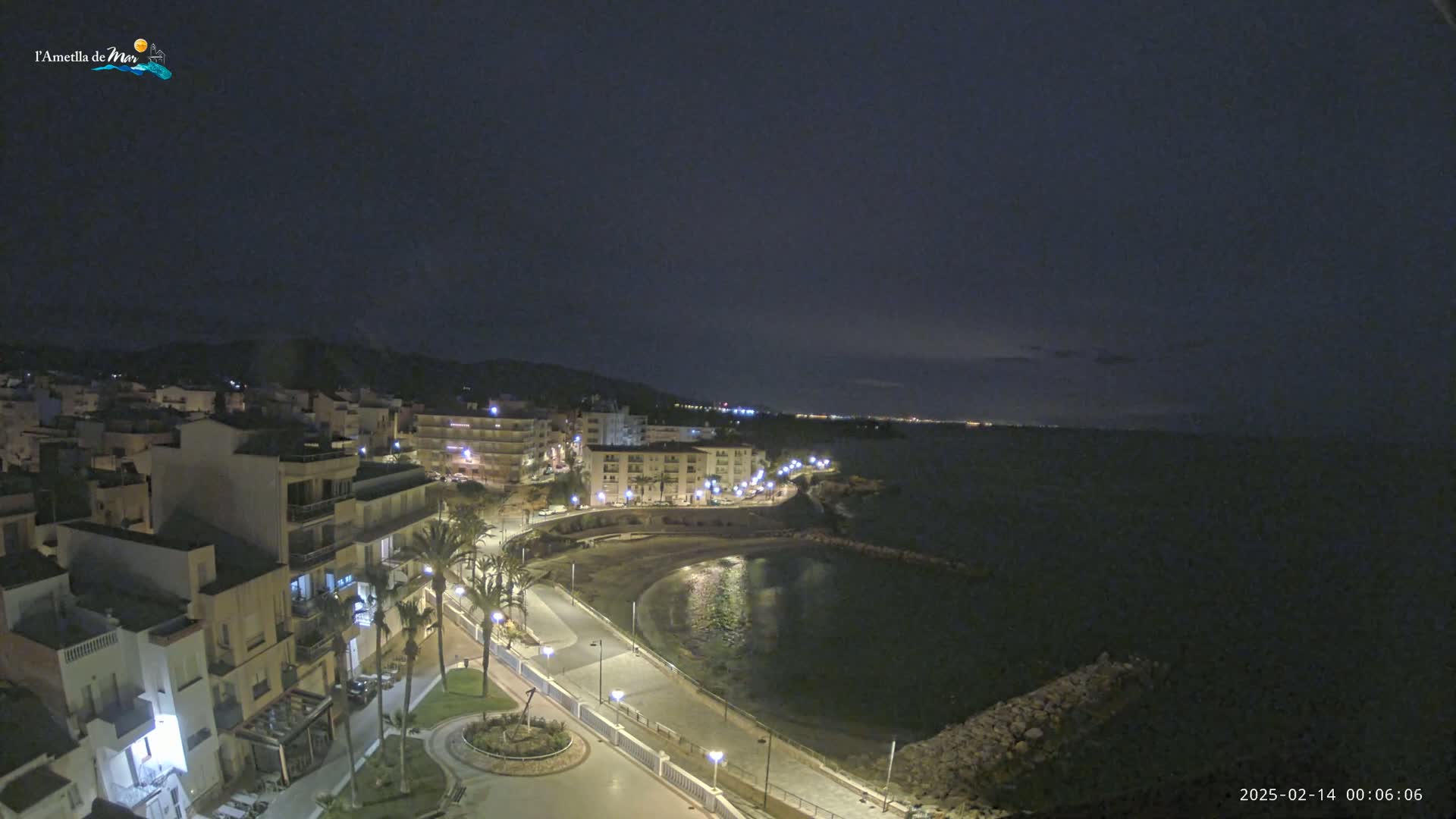 A nighttime coastal town scene shows a calm bay, a paved walkway lined with palm trees and streetlights, and multi-story buildings illuminated against a dark sky.