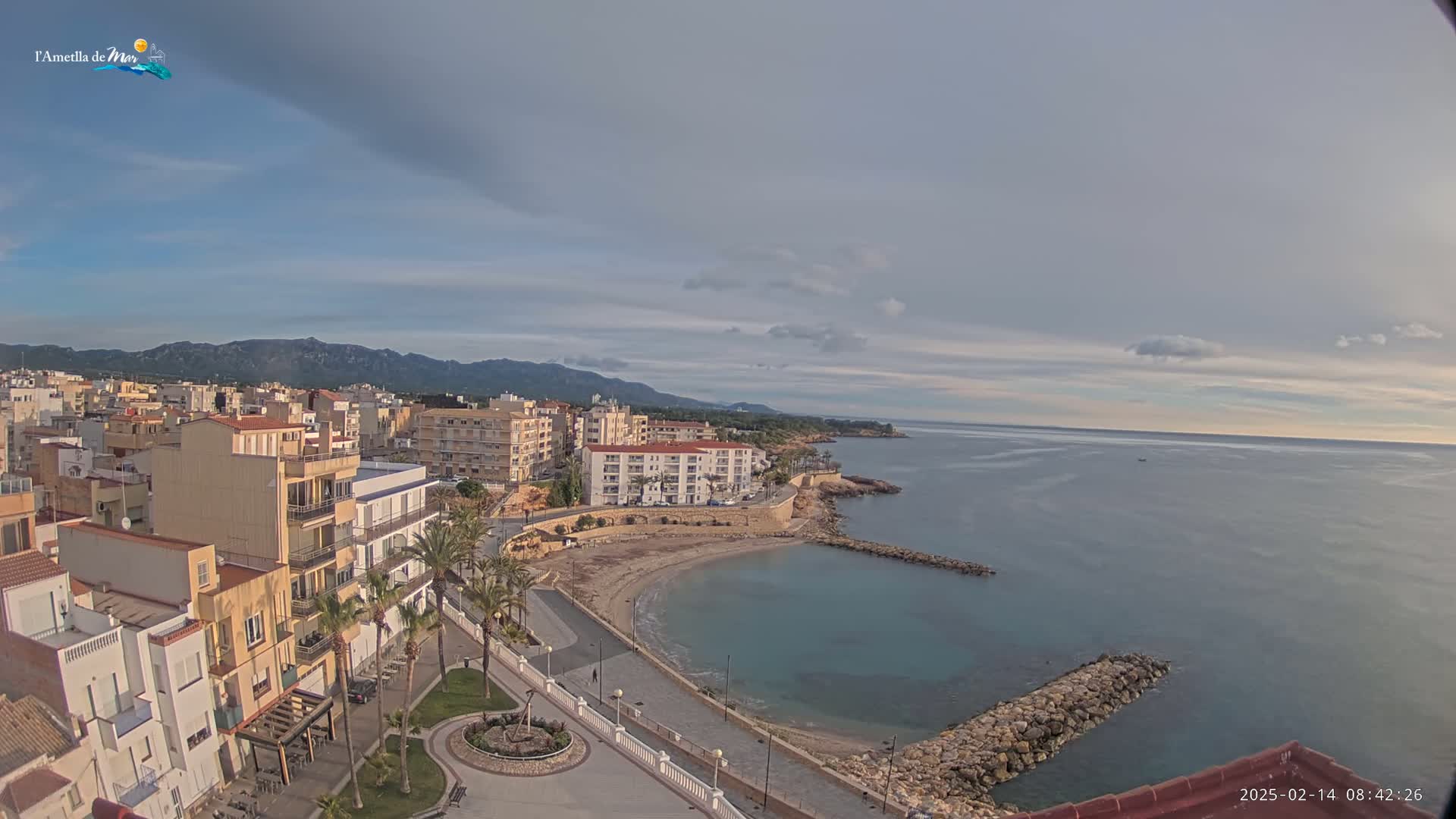 An aerial view shows a coastal town with buildings along a curved shoreline, a small sandy beach, and calm blue water under a partly cloudy sky.