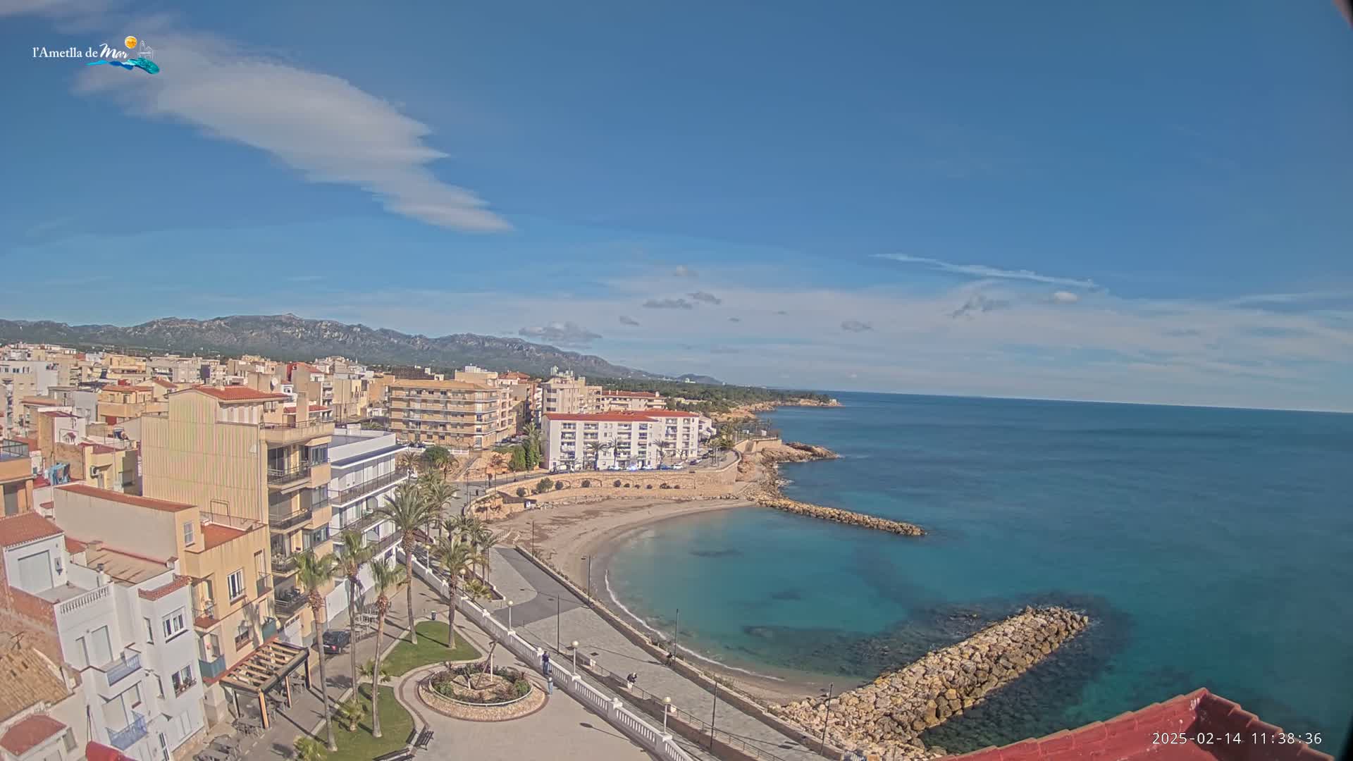A coastal town with buildings, a small sandy beach, and a clear blue sea under a mostly sunny sky.