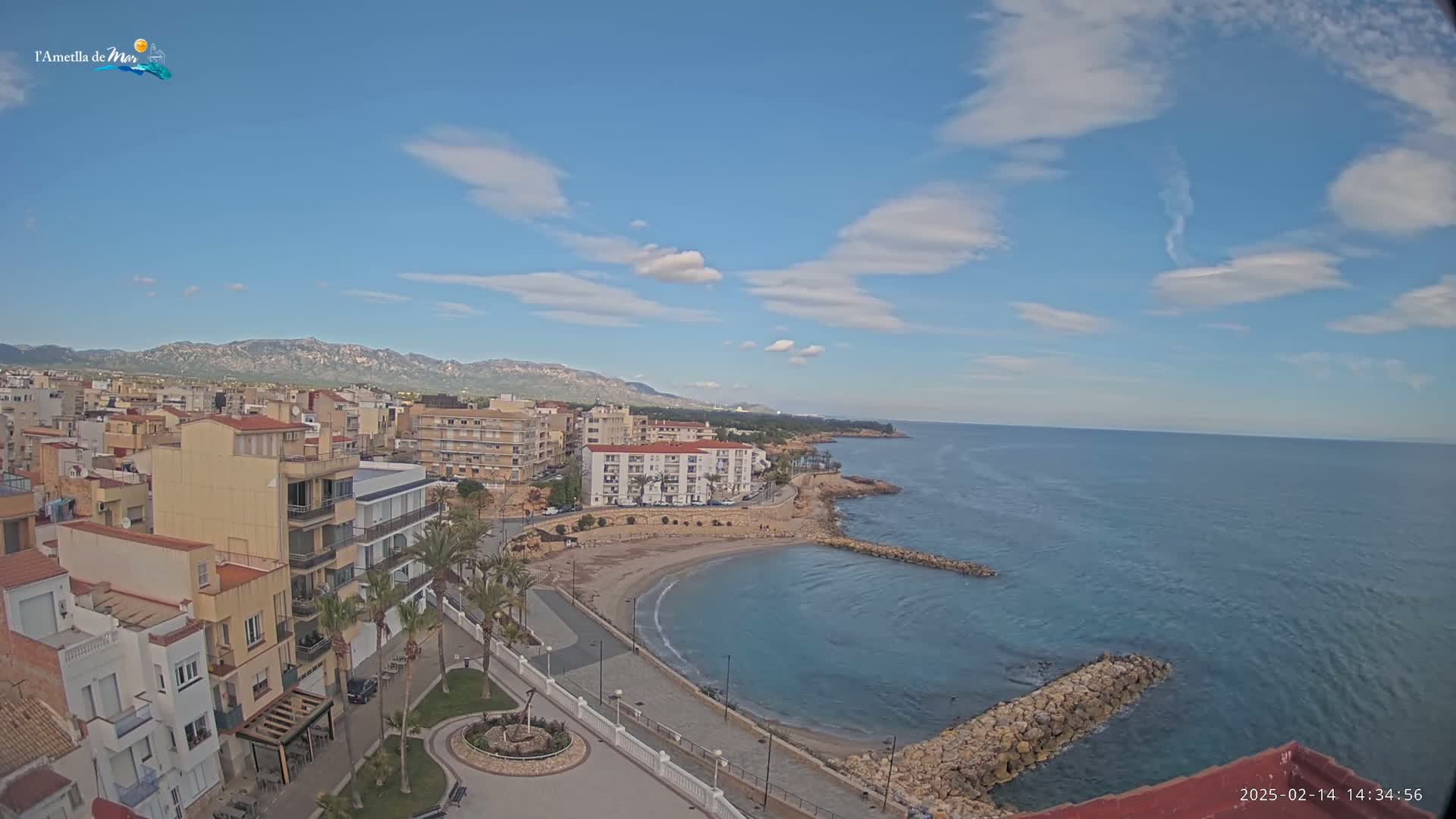 A coastal town with a small, sandy cove and calm, blue sea under a mostly sunny sky with scattered clouds.