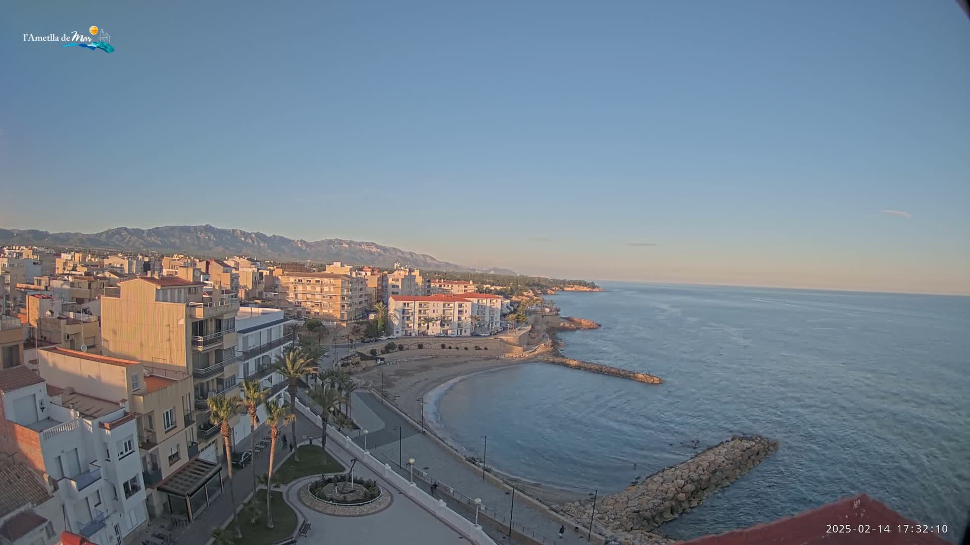 An aerial view shows a coastal town under a clear, pale blue sky, with calm water and a sandy beach curving into a small bay.