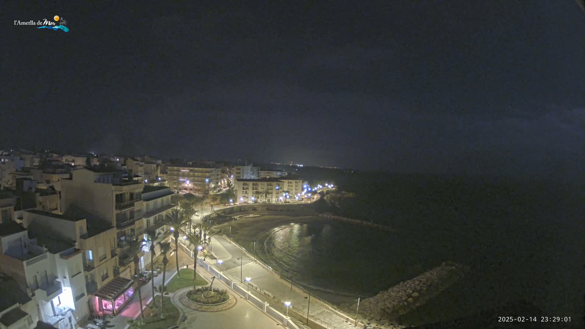 A nighttime, high-angle view shows a coastal town with buildings illuminated along a crescent-shaped bay under a dark sky.