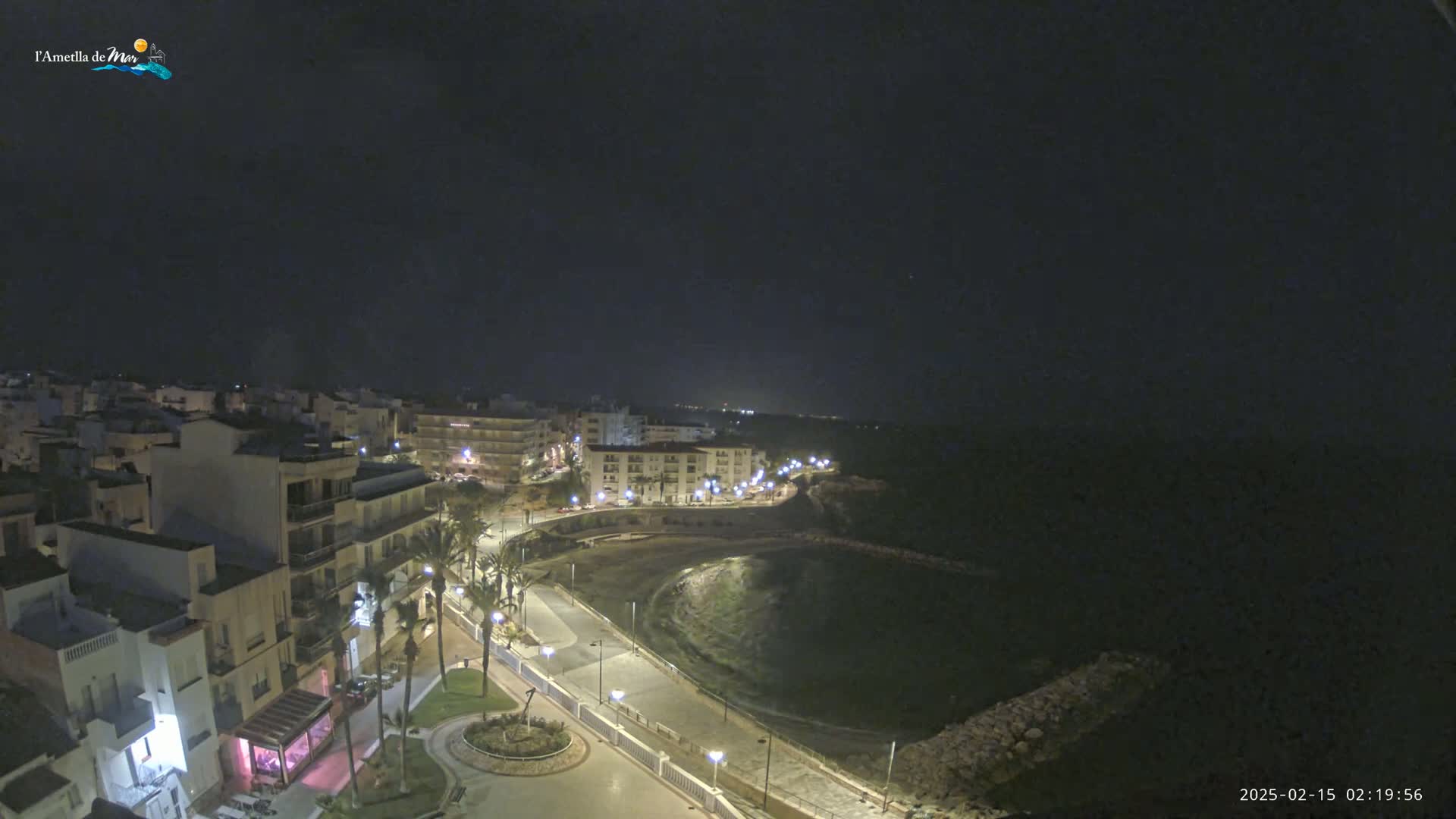 A nighttime aerial view shows a coastal town with buildings illuminated along a crescent-shaped bay under a clear, dark sky.