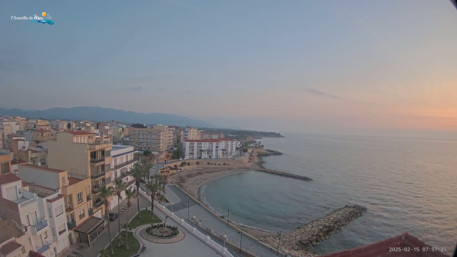 A coastal town at sunrise, with calm water and a small, crescent-shaped beach, is nestled against a backdrop of mountains.