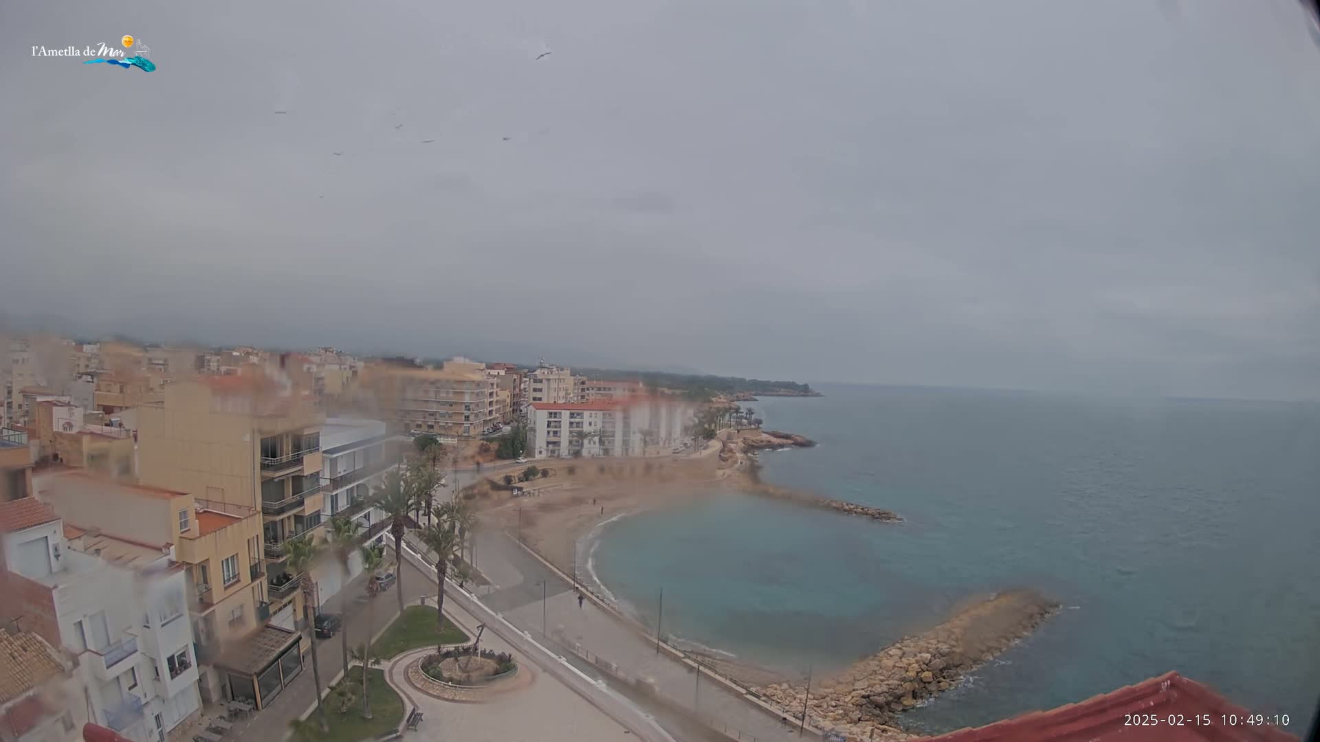 A coastal town is seen on a rainy day, with a small, calm bay and several multi-story buildings visible.