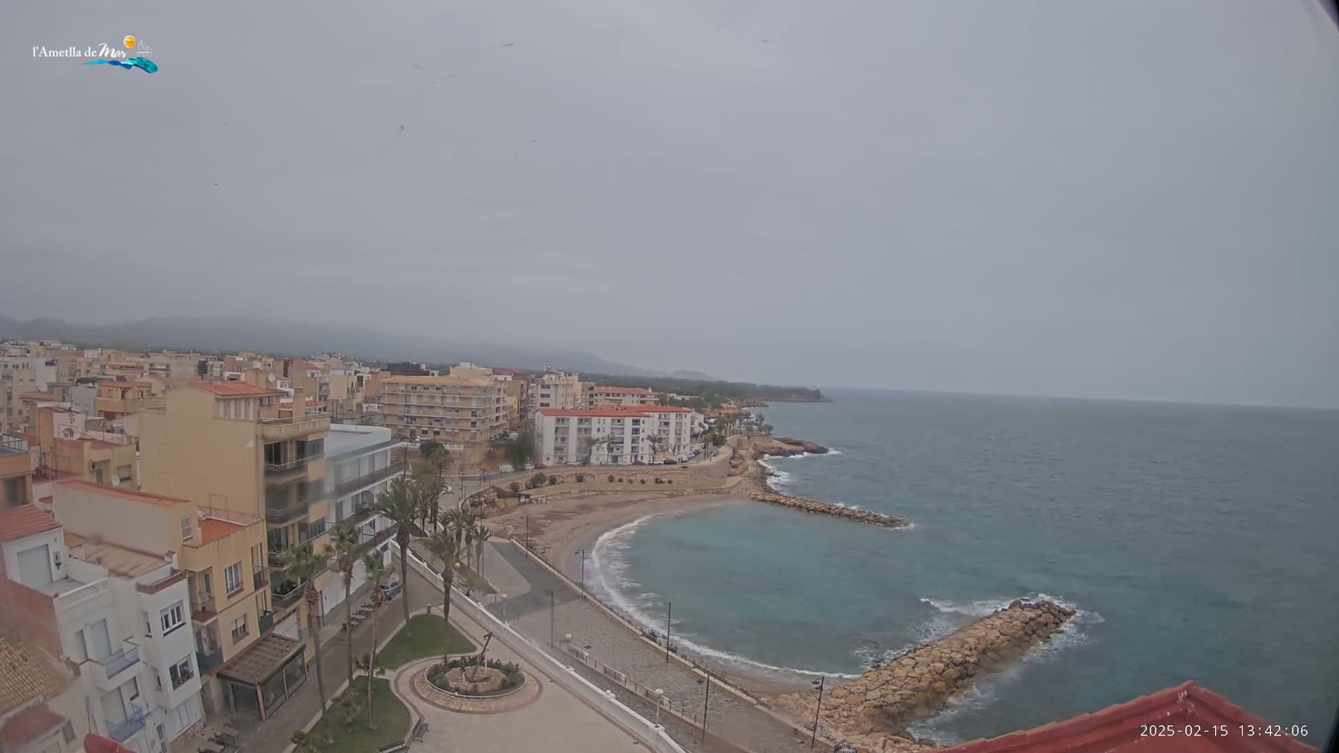An overcast day reveals a coastal town with buildings cascading down to a small, crescent-shaped beach and calm sea.
