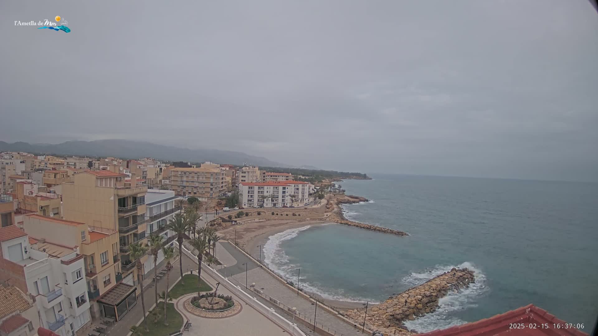 A coastal town with low-rise buildings sits on a grey, overcast day next to a calm, blue-green sea with small waves breaking against a rock jetty.