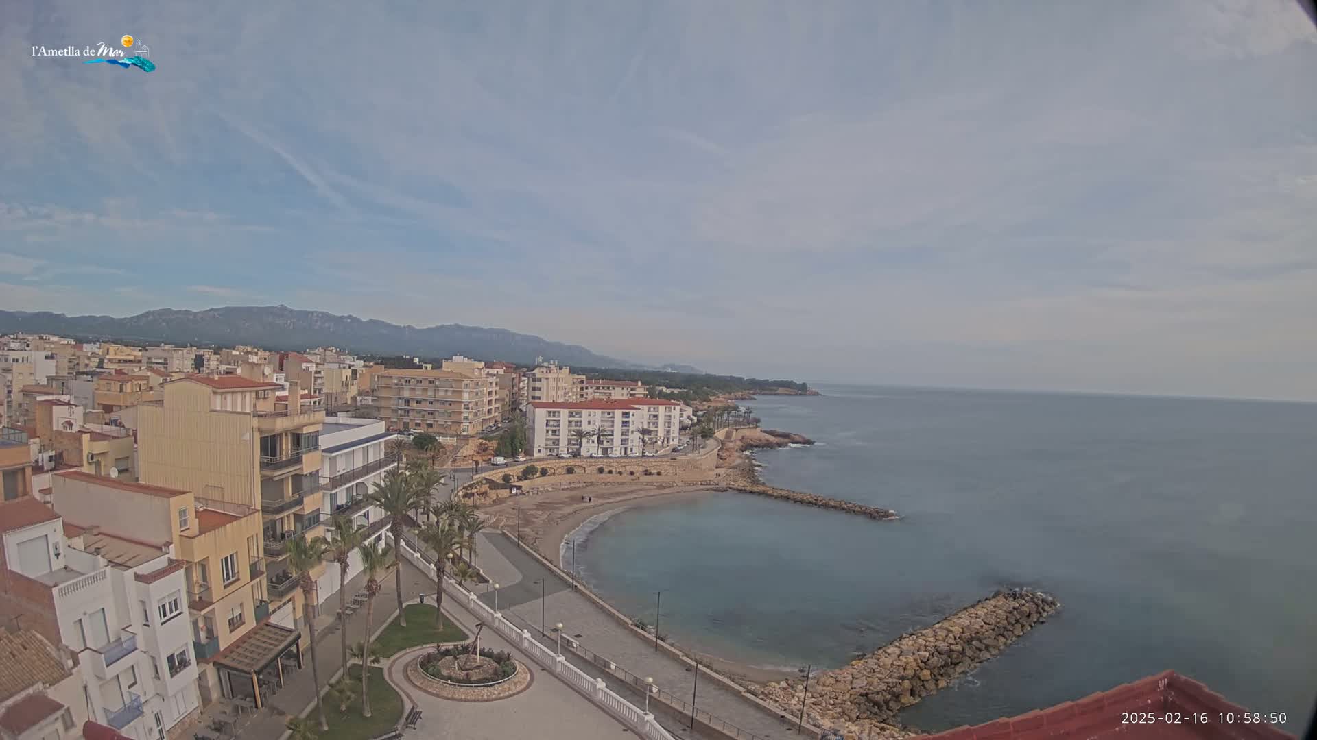 An aerial view shows a coastal town under a mostly clear sky, with a small sandy beach curving into a calm sea.