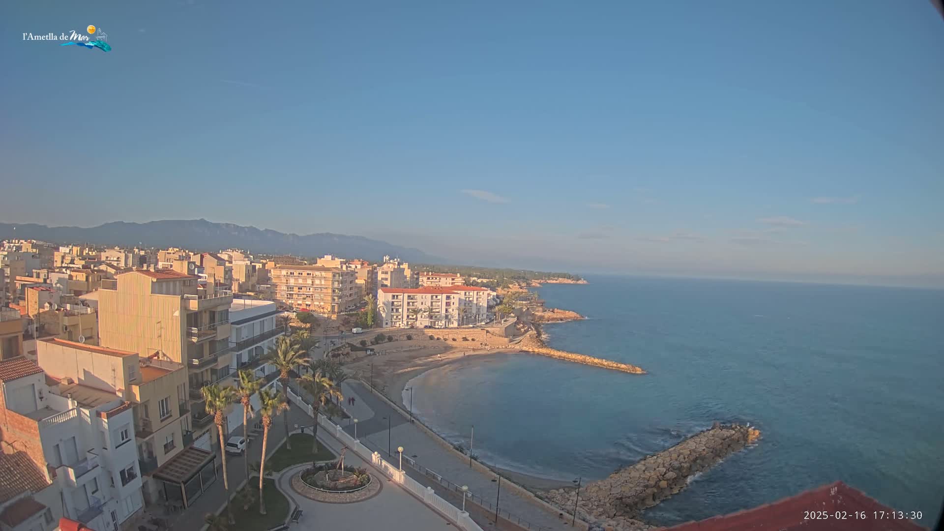 A coastal town with buildings, palm trees, and a small beach sits on a calm, clear sea under a mostly clear sky, with mountains visible in the distance.