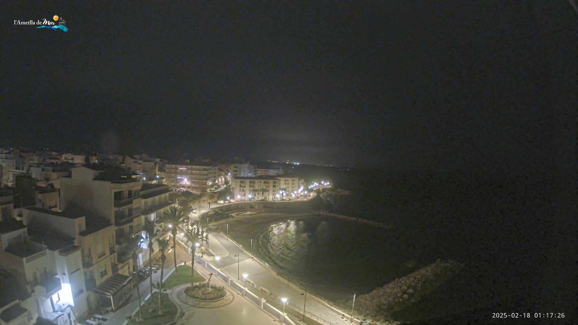 A nighttime, high-angle view shows a coastal town with buildings lit along a paved waterfront path curving around a dark, calm bay.
