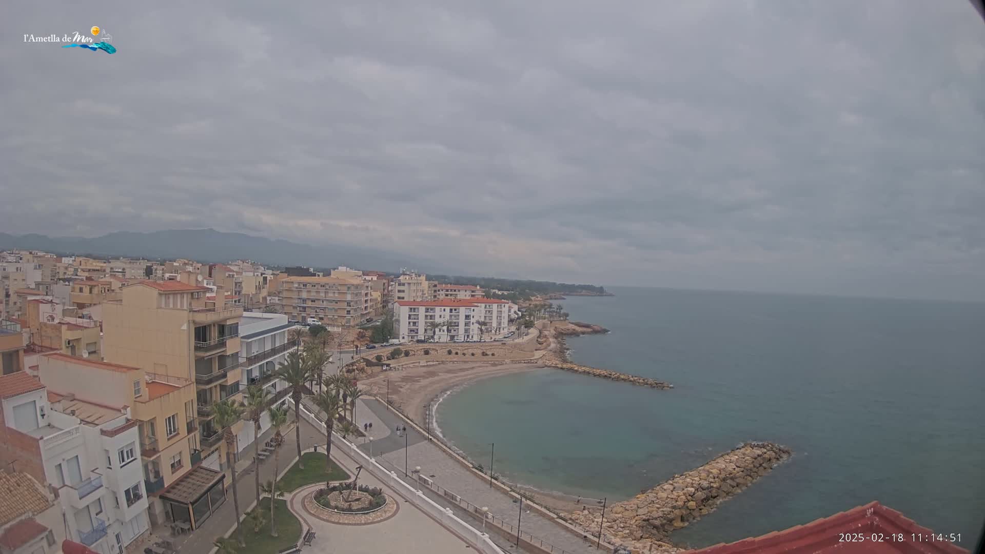 An overcast day reveals a coastal town with buildings, a small sandy cove, and a calm sea extending to distant mountains.