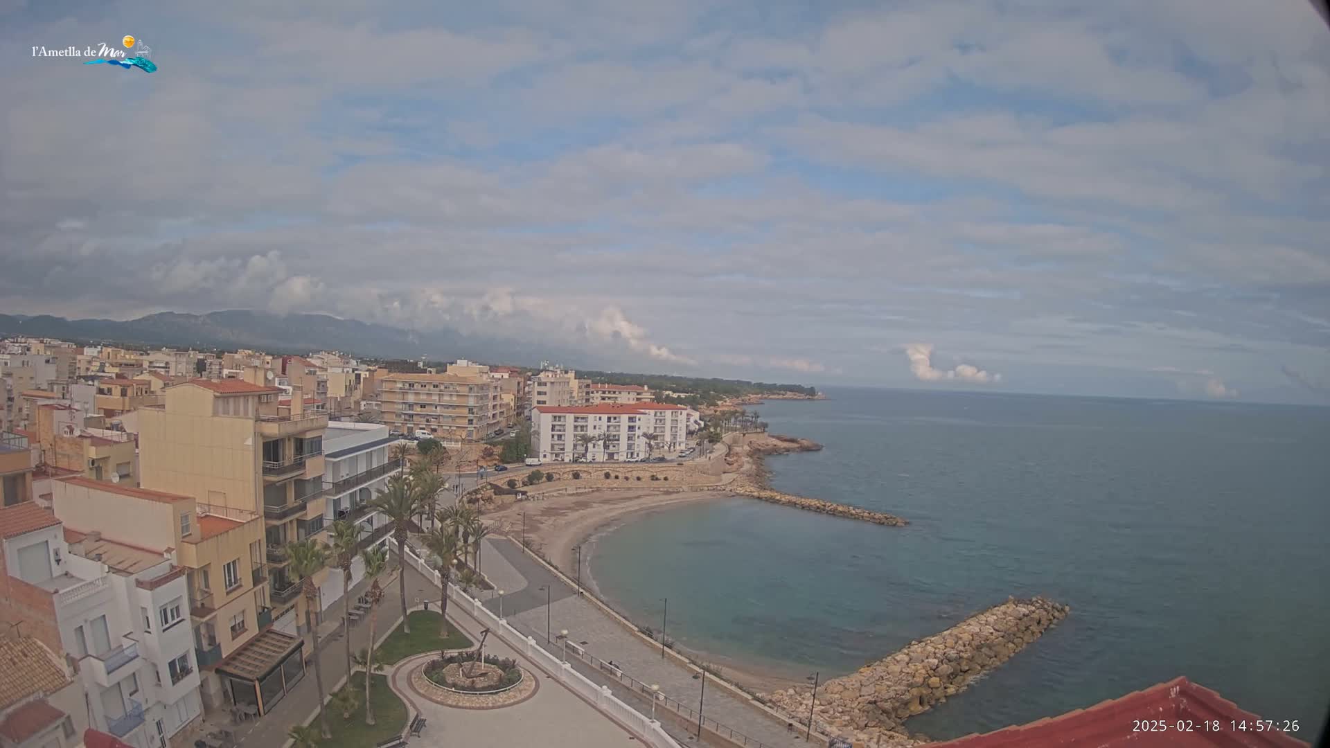An aerial view shows a coastal town on a partly cloudy day, with a small sandy cove and calm ocean water.