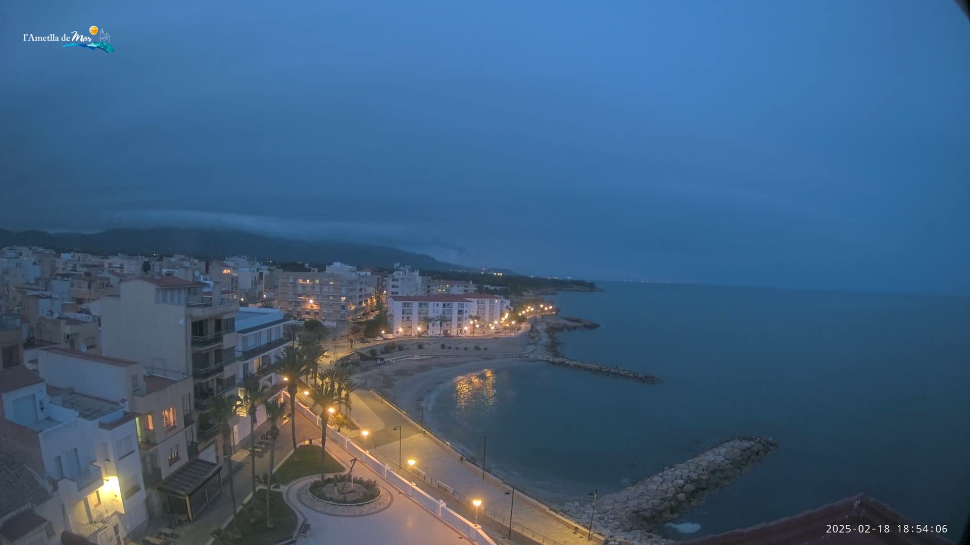 A coastal town at dusk, with a calm sea under a mostly cloudy sky, is illuminated by streetlights along a paved waterfront path.