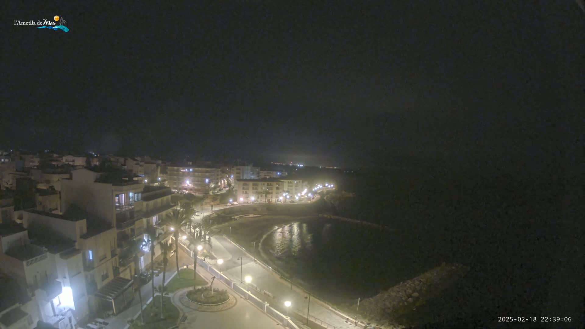 A nighttime, high-angle view of a coastal town with illuminated buildings and streets curving alongside a dark body of water.
