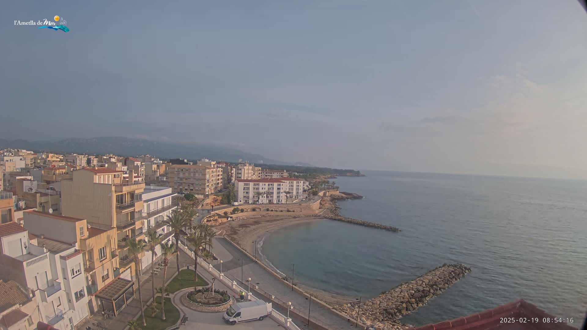 A coastal town with multi-story buildings is situated next to a calm, blue sea under a hazy sky.