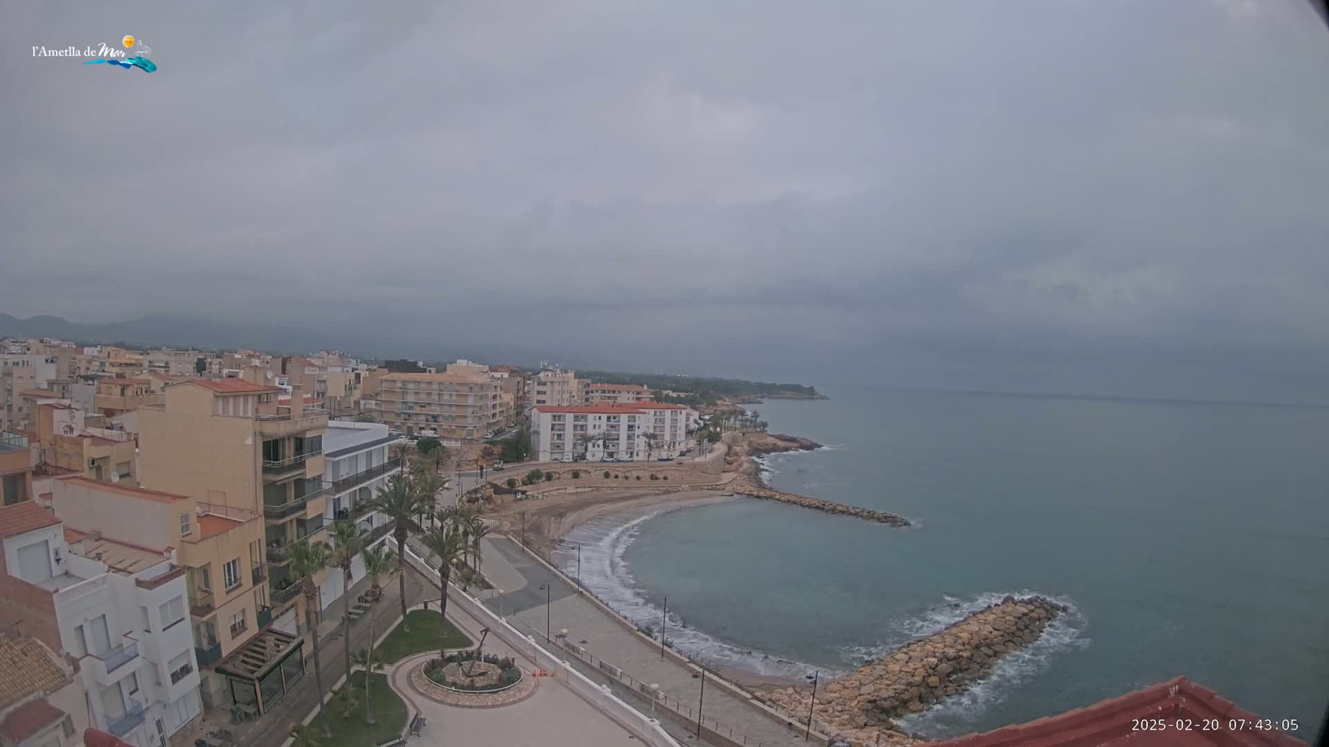 An overcast day reveals a coastal town with low-rise buildings, a sandy beach, and a calm sea partially protected by a rock breakwater.