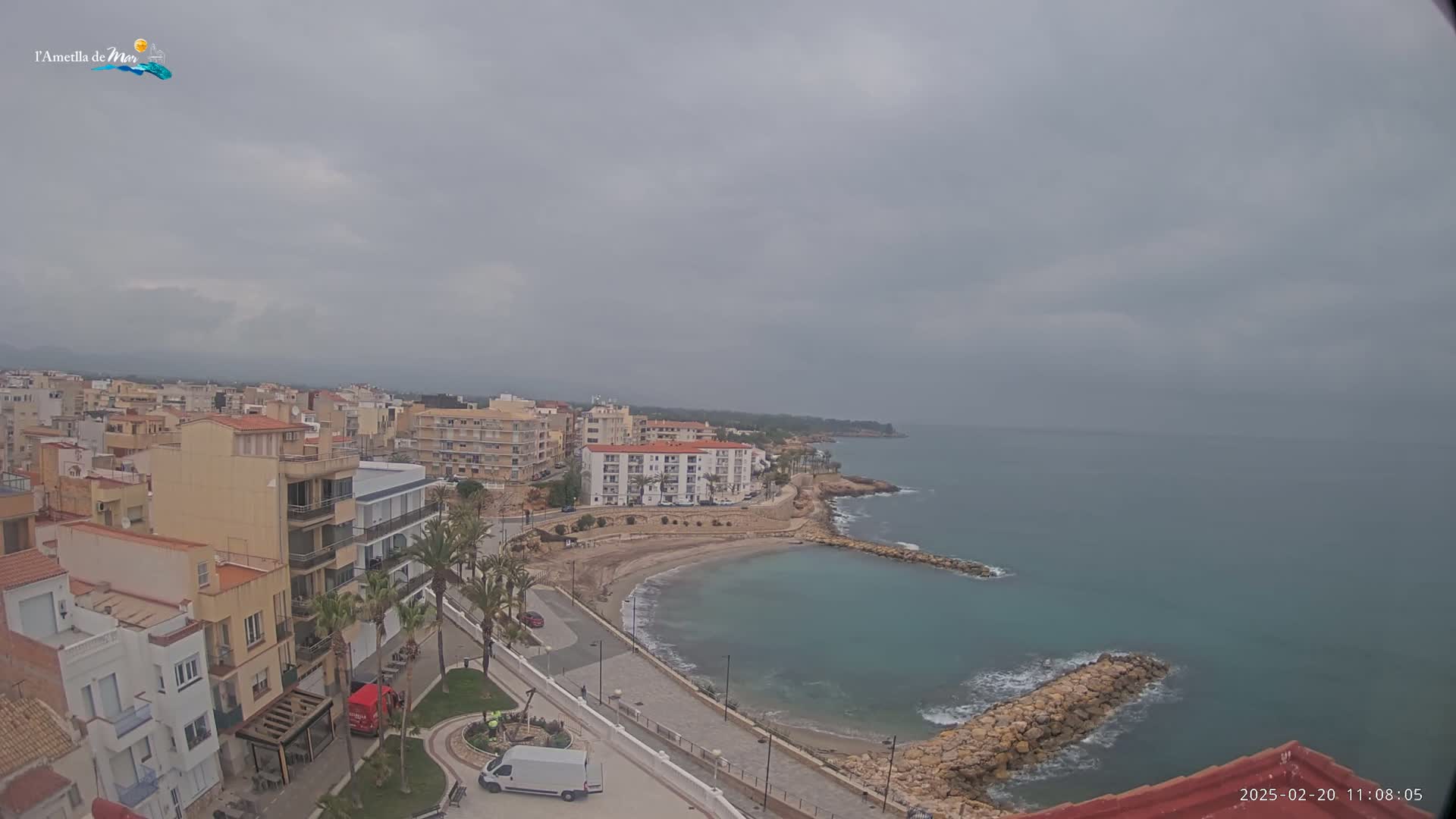 An elevated view of a coastal town on an overcast day shows buildings lining a small, calm bay with a rocky breakwater, and a sandy beach.