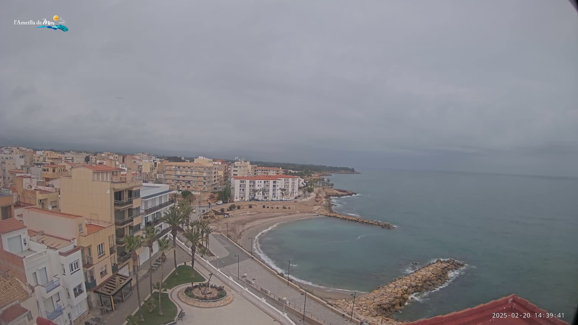 An overcast day reveals a coastal town with buildings, a small sandy cove, and calm, blue-green water.