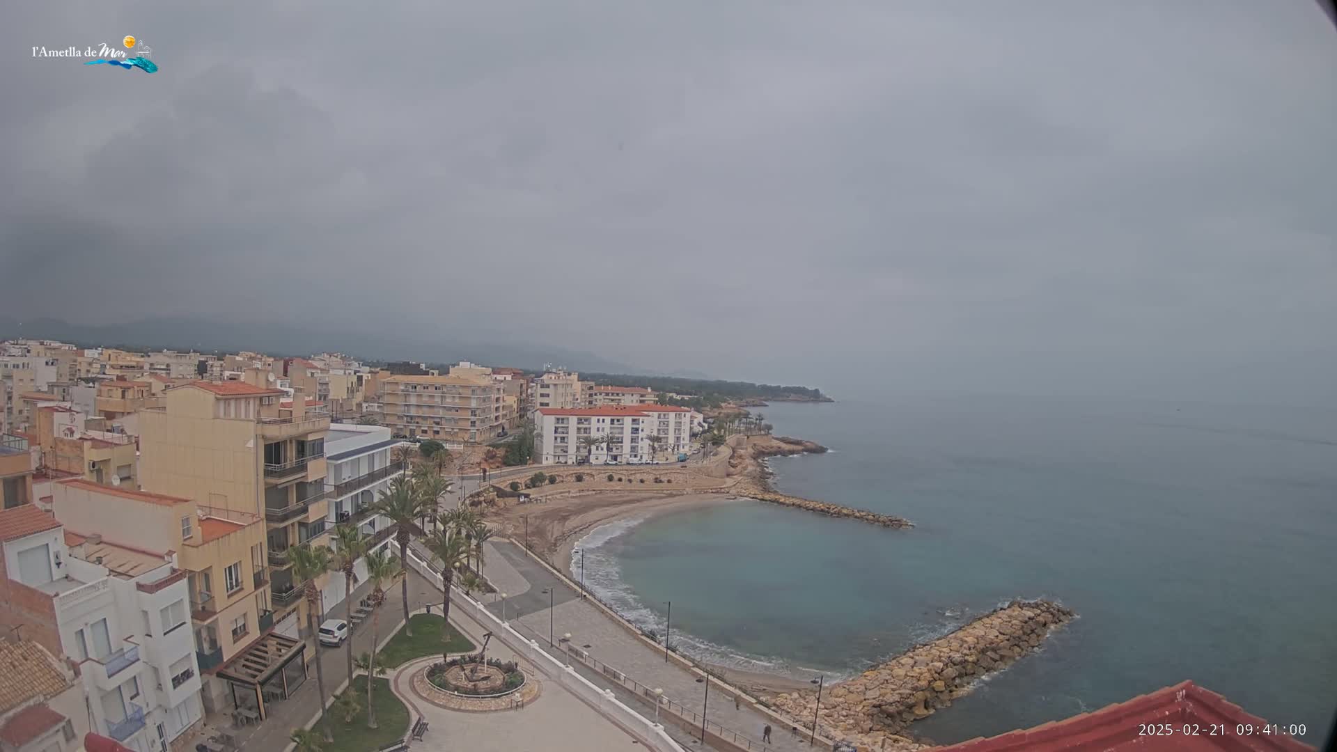 An overcast coastal town with buildings, a small sandy cove, and a rocky breakwater extends into a calm sea.