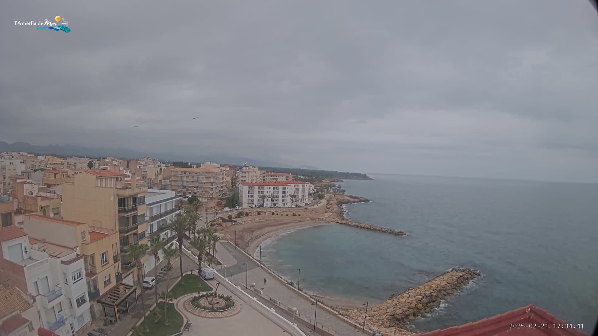 An overcast day reveals a coastal town with buildings lining a small, sandy cove that opens to a calm sea.