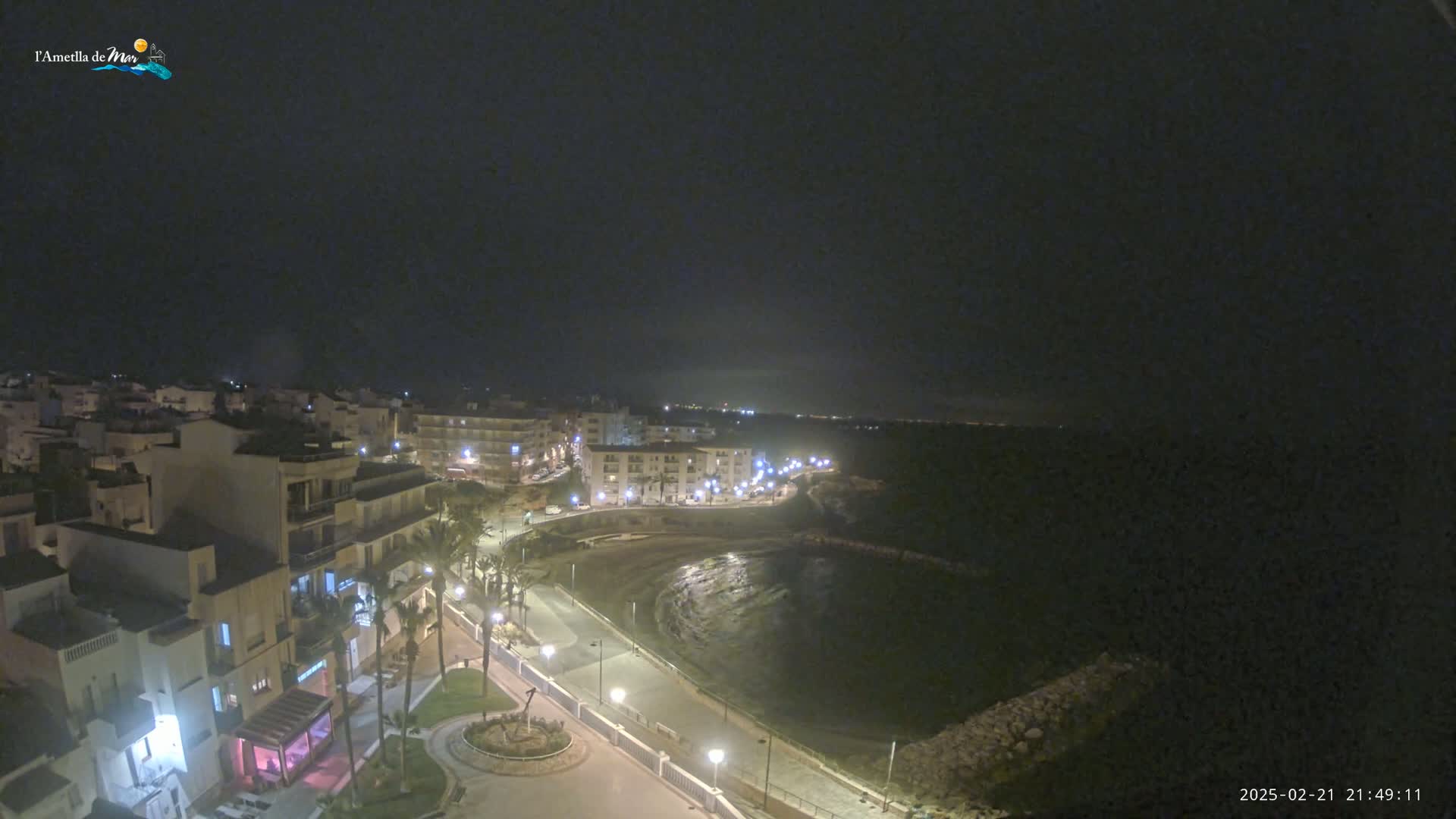 A nighttime, high-angle view shows a coastal town with a calm bay, illuminated by streetlights.