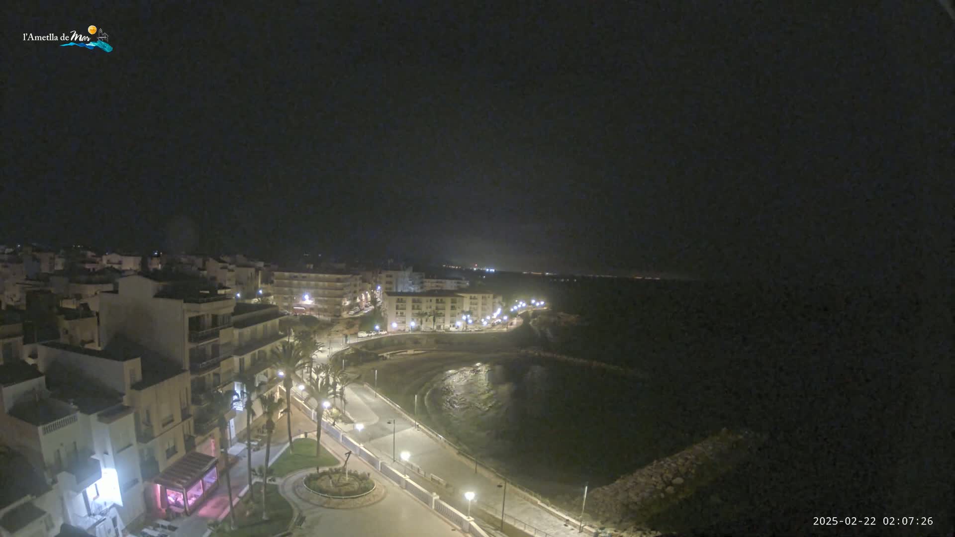 A nighttime, high-angle view of a coastal town with a calm bay, featuring buildings lit with lights along a curved road.