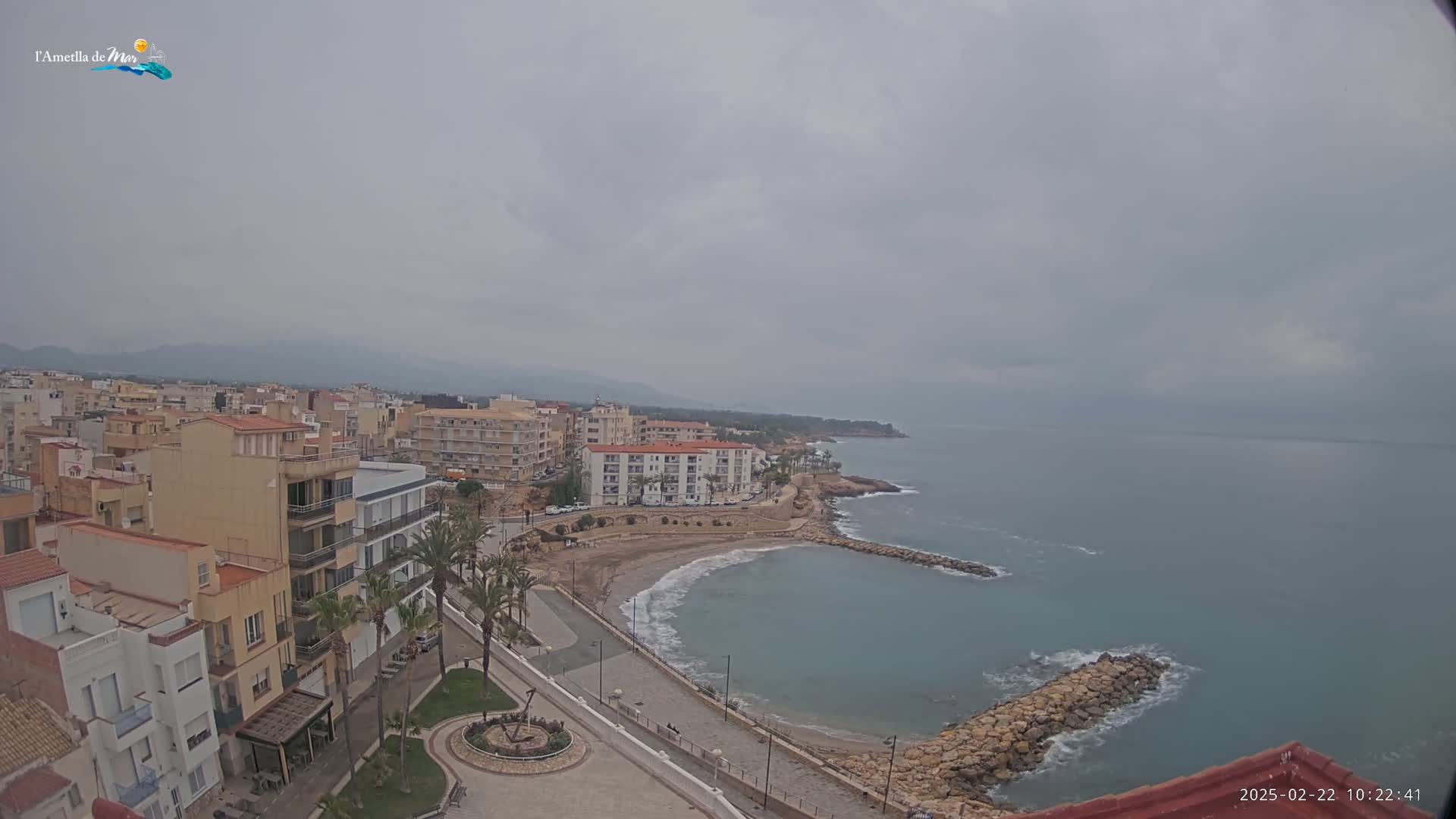 A coastal town with multi-story buildings is seen on an overcast day, with a calm sea and a small sandy cove visible in the foreground.