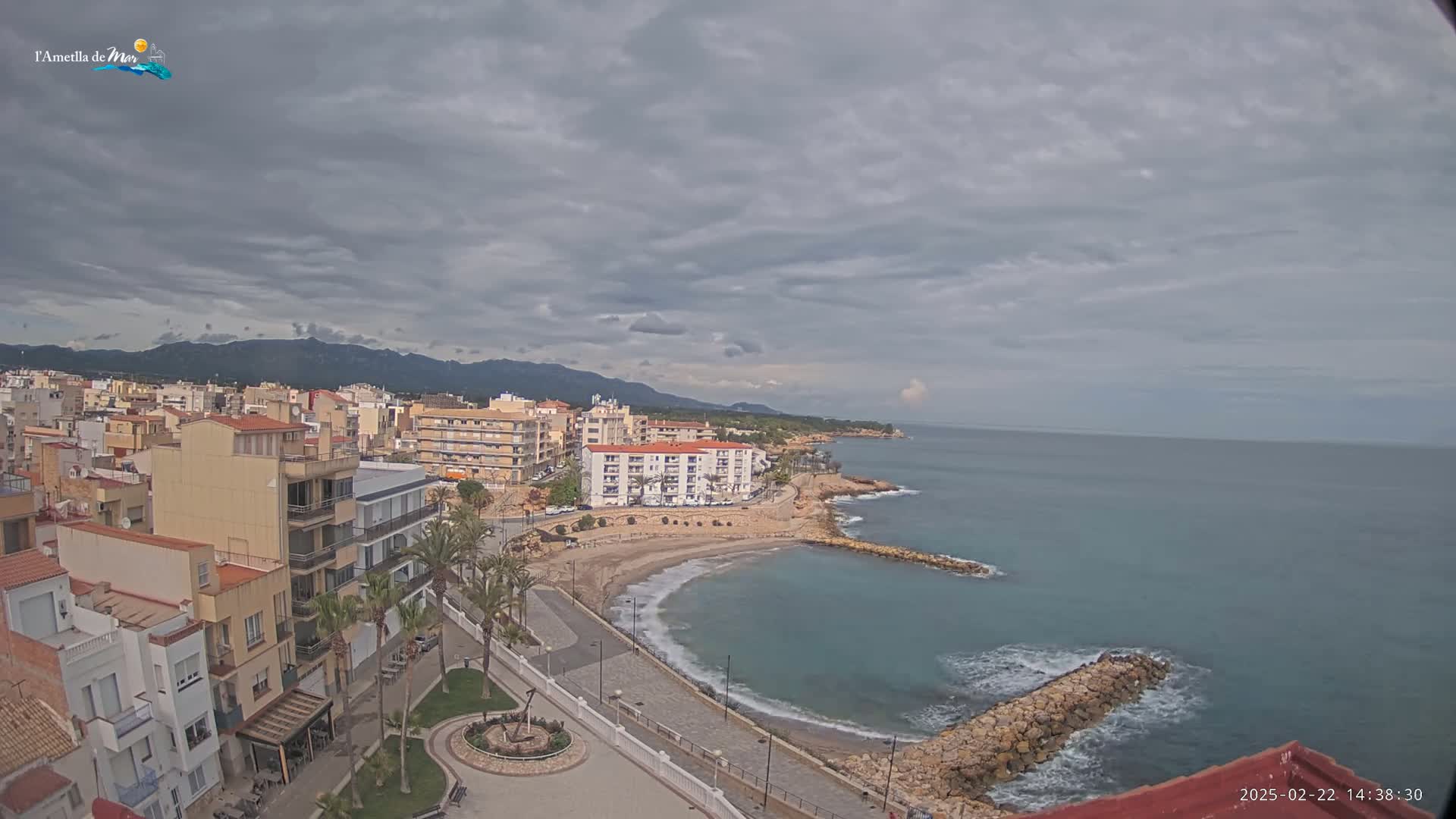 An aerial view shows a coastal town under a cloudy sky, with a calm sea lapping a small sandy beach.