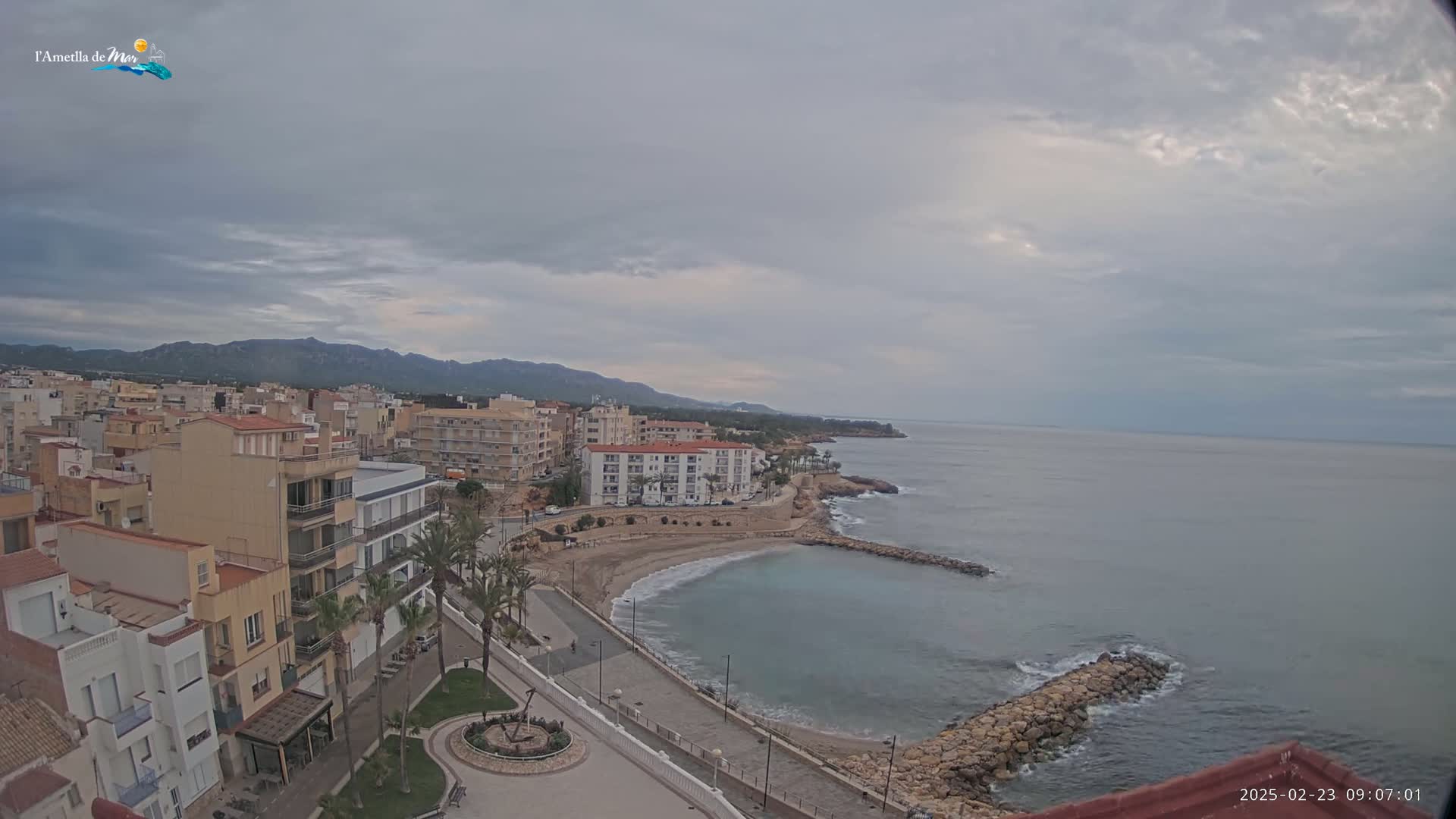 An overcast day reveals a coastal town with buildings, palm trees, a small beach, and a calm sea stretching to distant mountains.