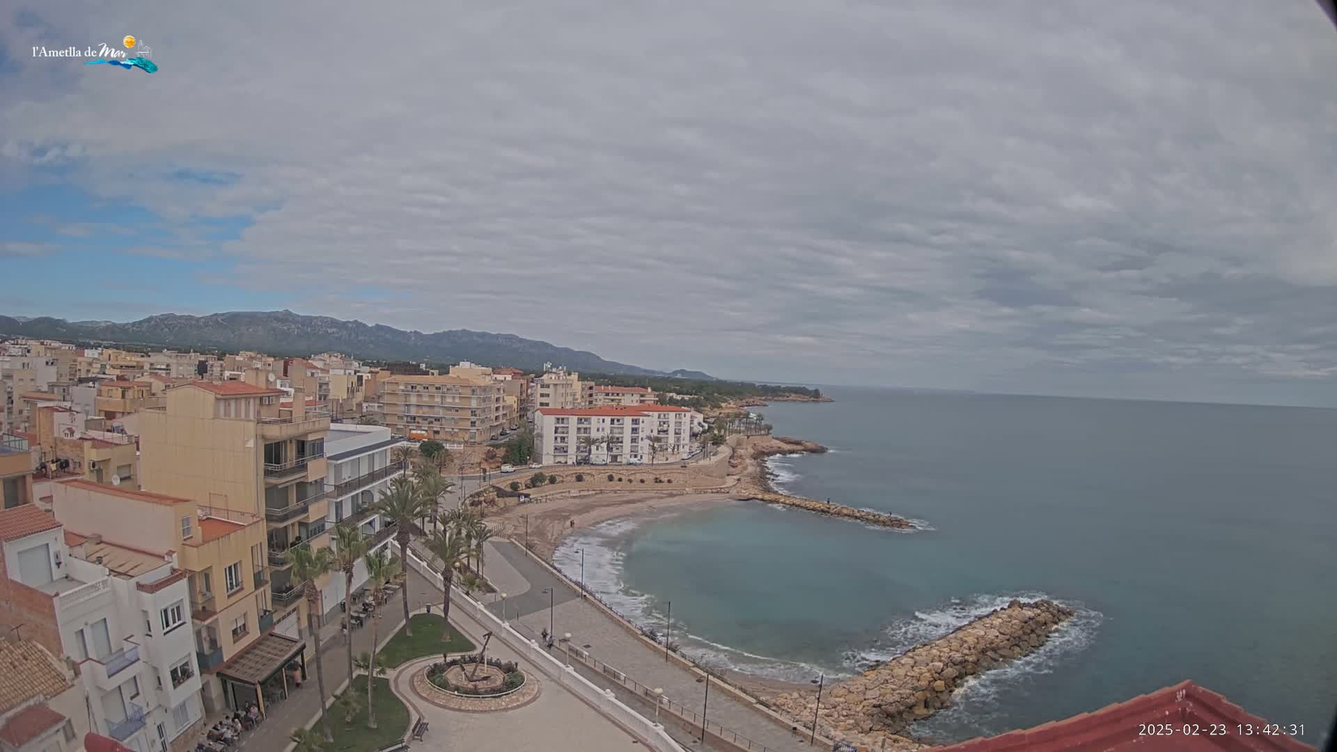 An elevated view shows a coastal town under a cloudy sky, with a calm sea lapping a small, sandy cove next to a rocky shoreline and buildings stretching inland toward distant mountains.