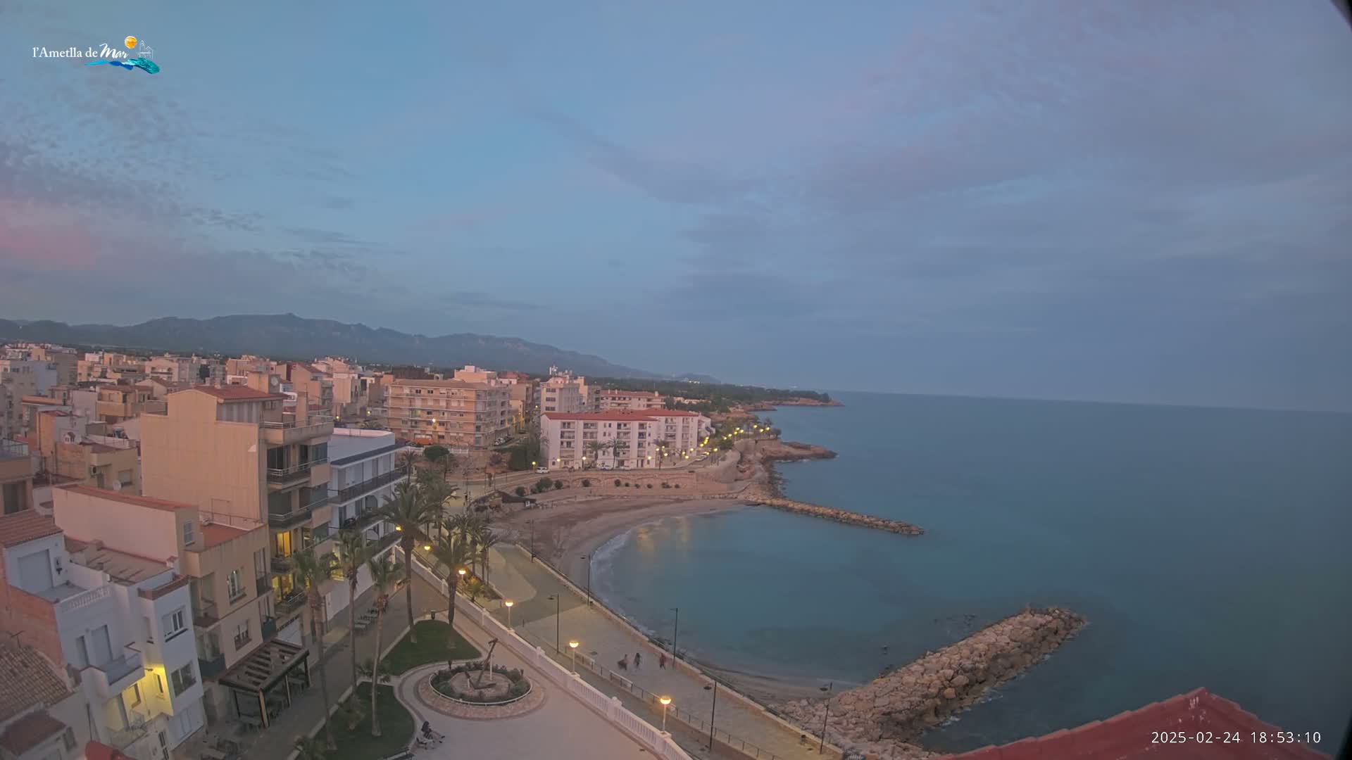 An elevated view shows a coastal town at dusk under a mostly clear sky, featuring buildings along a curving shoreline with a calm sea.